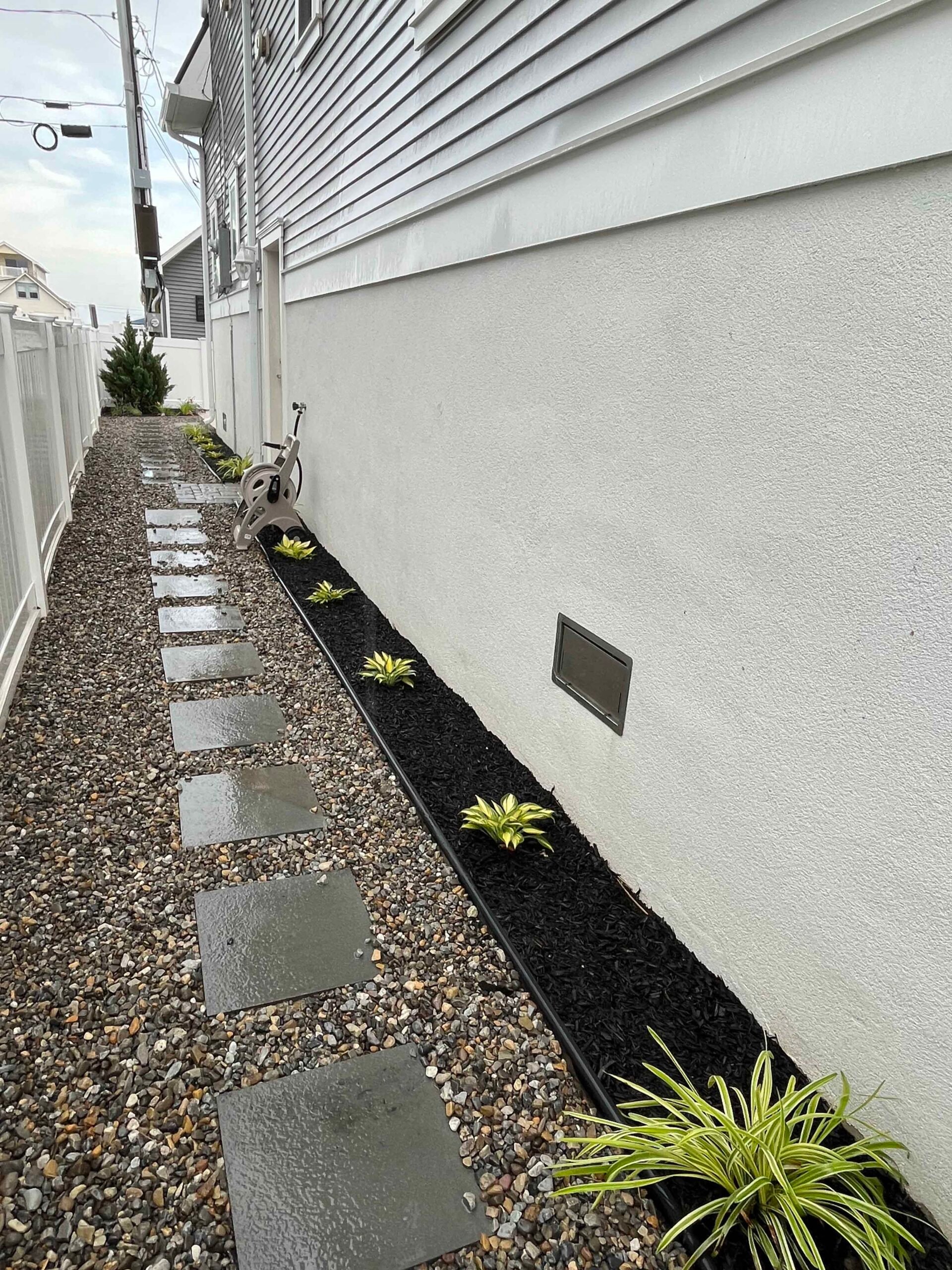 A narrow side-yard walkway features evenly spaced square bluestone stepping stones set into pea gravel, running alongside a stucco wall. A narrow planting bed along the wall is filled with black mulch and young ornamental grass or agave-type plants with ye