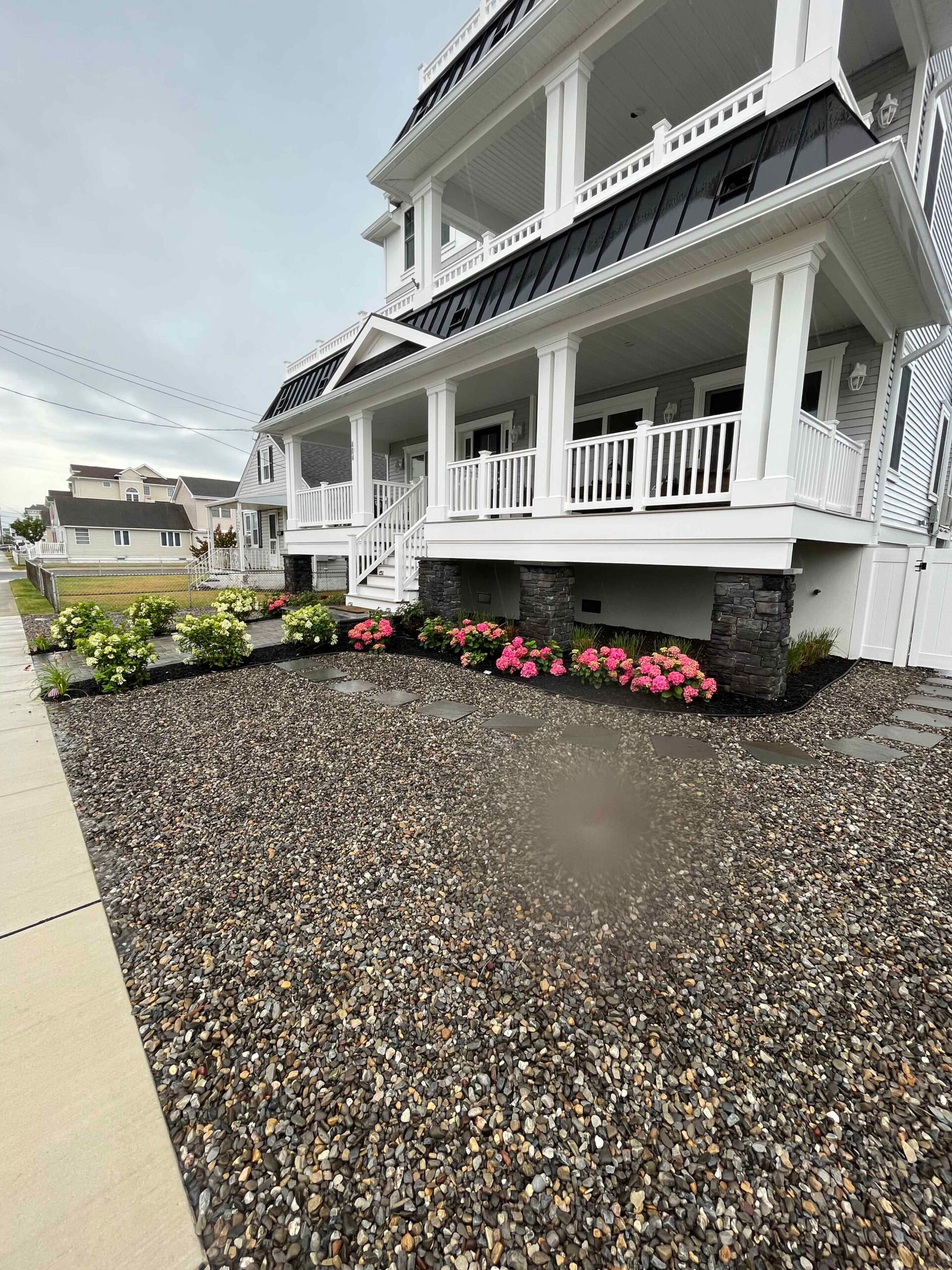 Pink and white hydrangea shrubs are planted in dark-mulched foundation beds beneath a multi-story coastal home's covered porch, with a large river rock and mixed pebble ground cover filling the front yard in place of lawn.