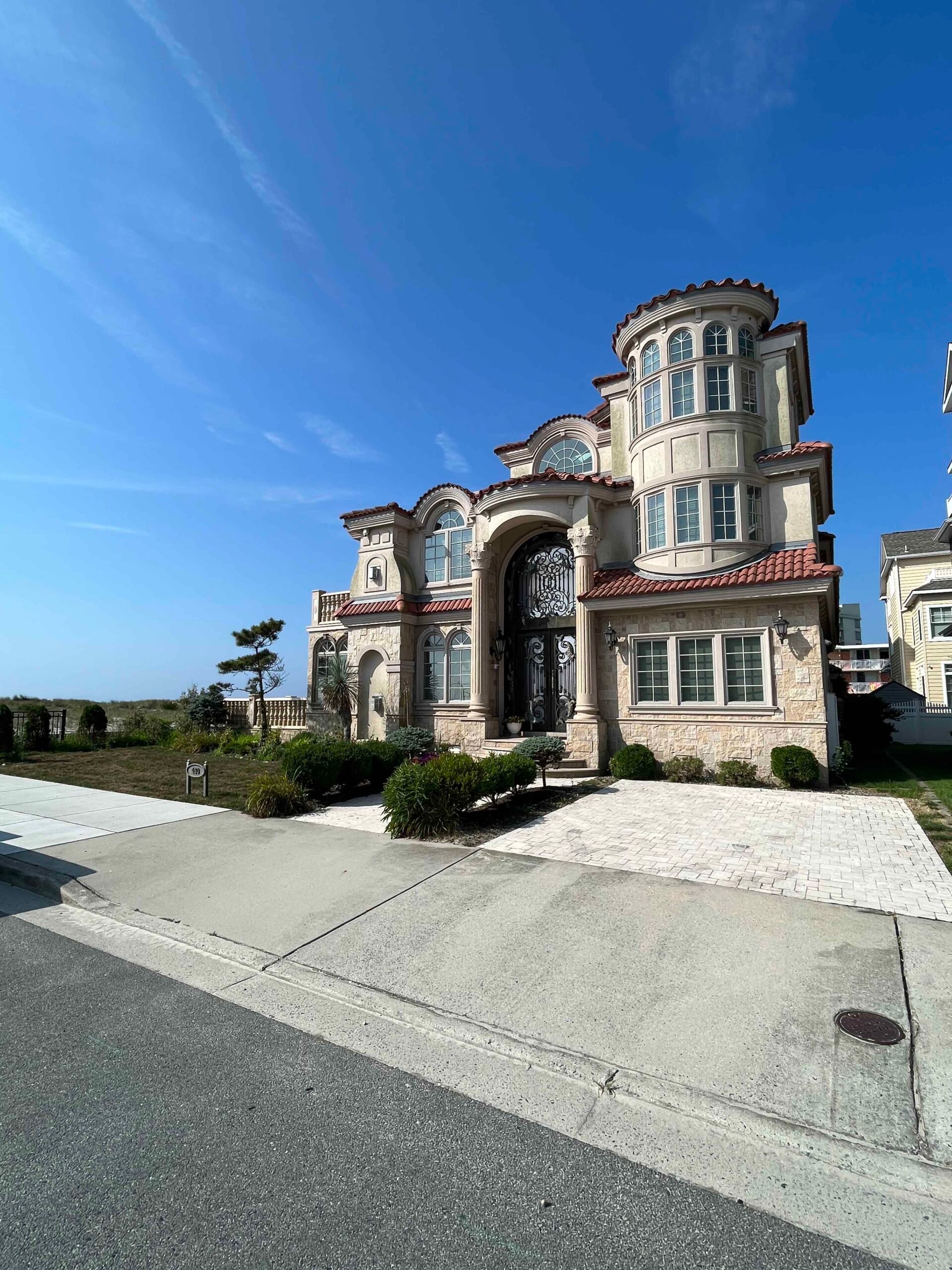 A large Mediterranean-style stucco and stone home features a concrete driveway with a decorative light-colored paver apron at the entrance. Low rounded boxwood-style shrubs are planted along the front foundation on either side of the ornamental iron entry 