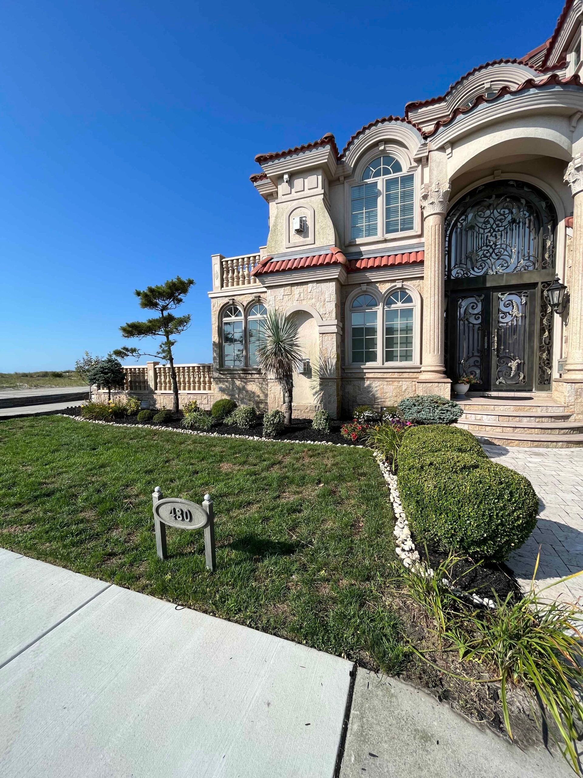 The front facade of a large estate home is framed by shaped boxwood spheres, ornamental grasses, yucca plants, and a small sculptural pine in beds filled with white decorative stone mulch. Beds follow the curve of a concrete sidewalk leading to an ornate w