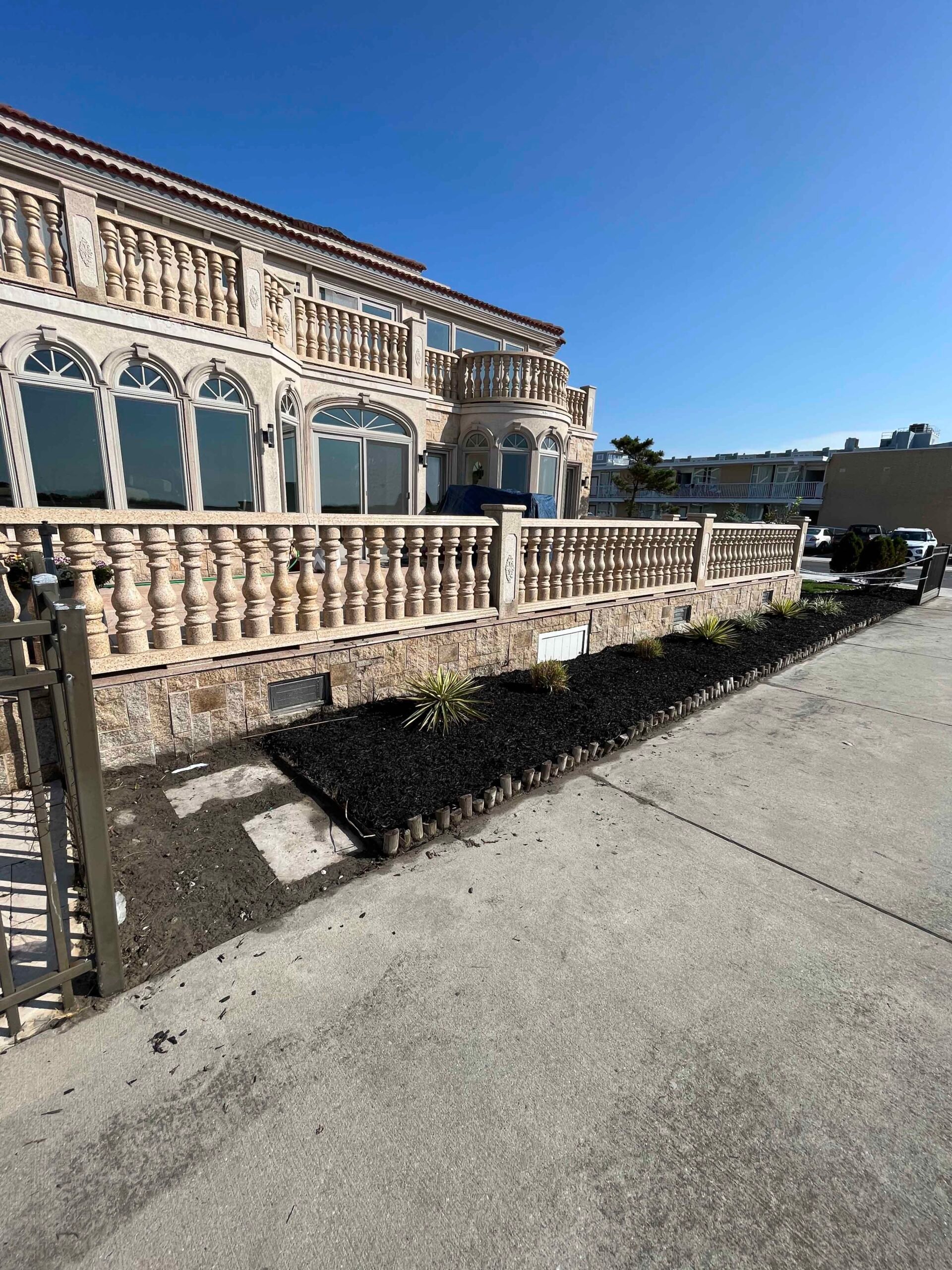 A wide planting bed runs along the front of a Mediterranean-style masonry building, freshly mulched with black mulch and edged with a row of short log-style timber edging. Several young ornamental plants, including what appears to be a spiky yucca or cordy