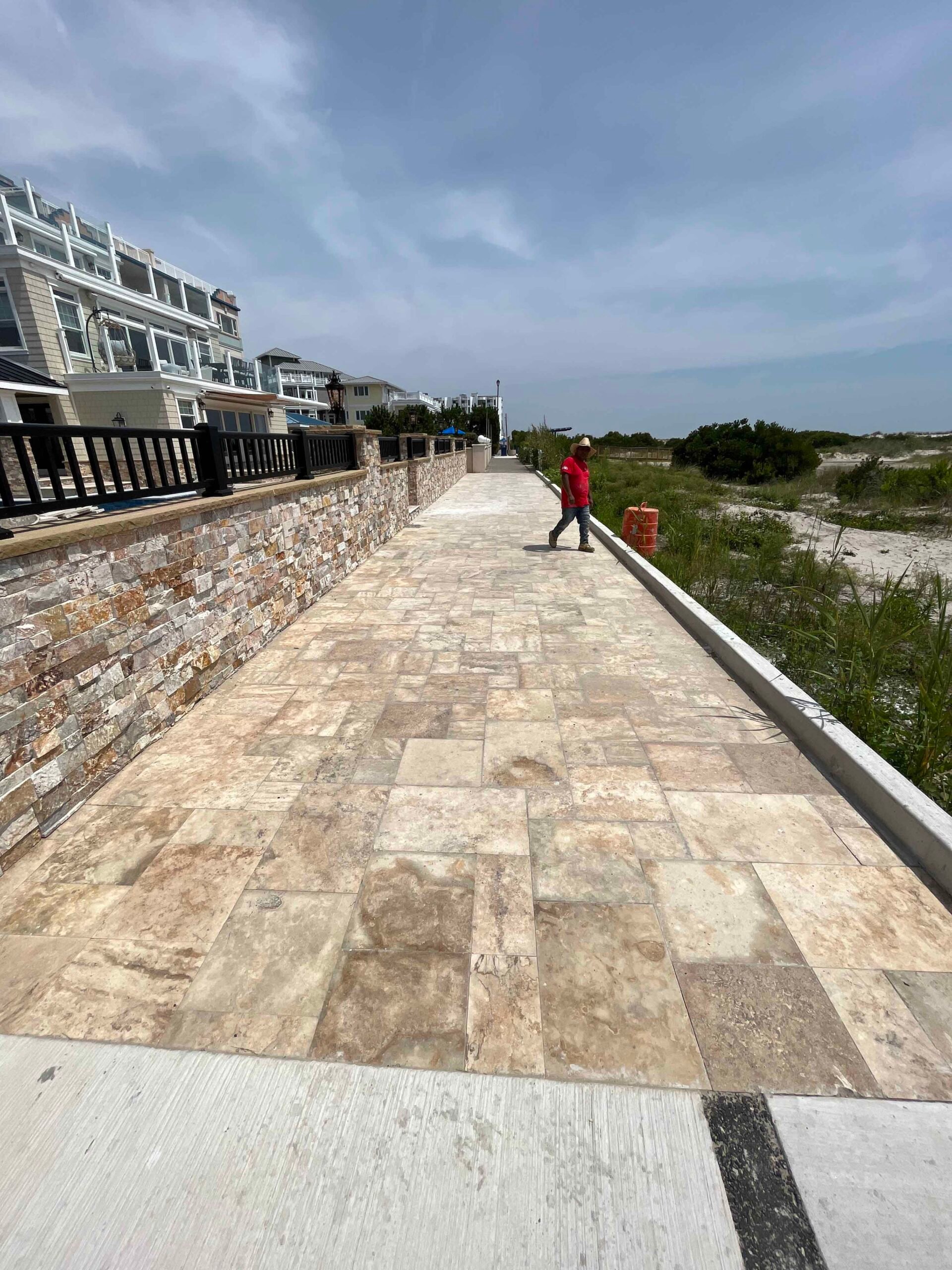 A wide travertine paver walkway runs along a natural stone stacked retaining wall beside a coastal property, with native dune grasses and open beach visible on the opposite side. A worker in a red shirt walks the length of the newly installed path.