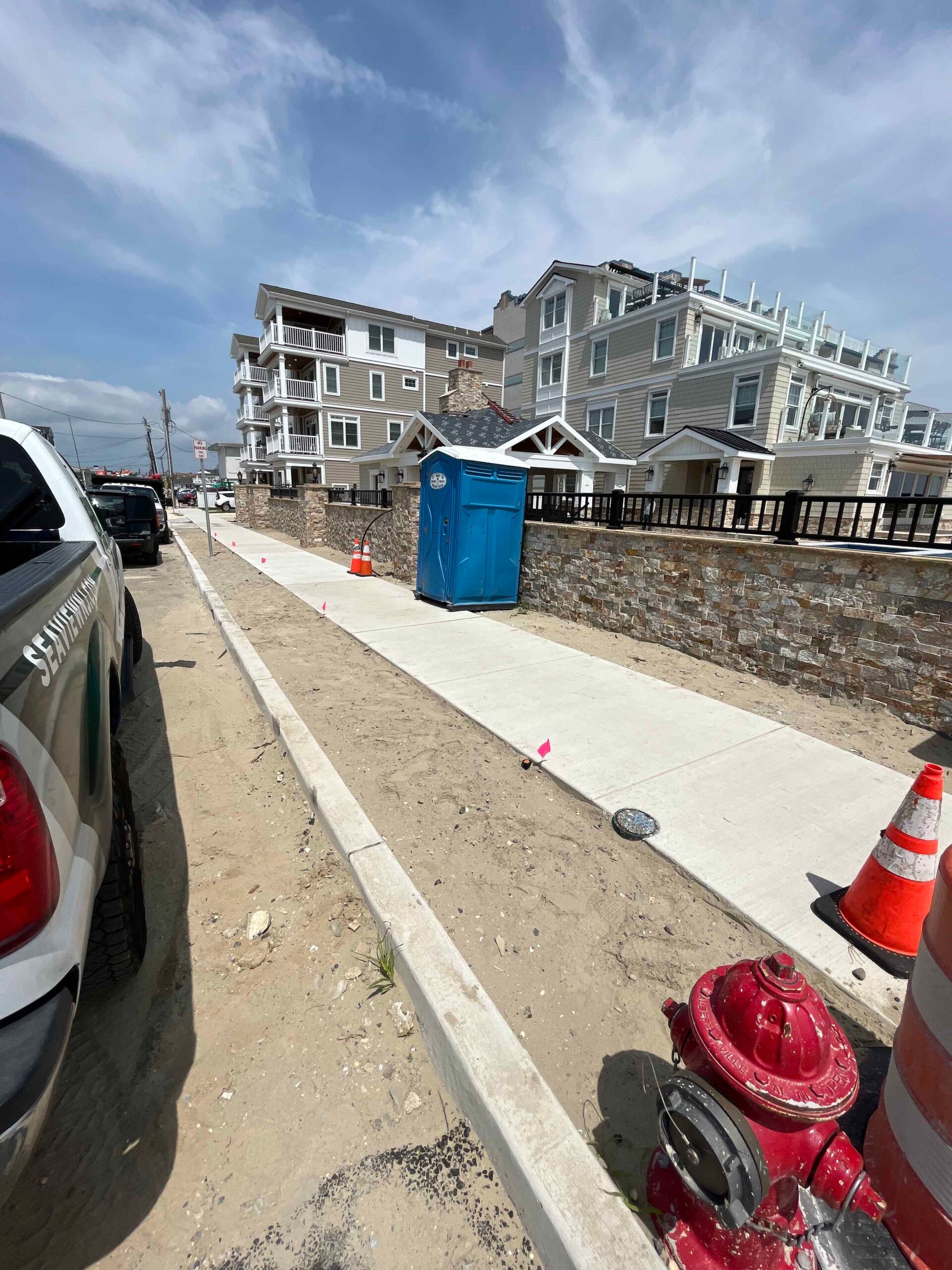 Freshly poured concrete sidewalk and curbing run along a coastal street in front of a newly constructed natural stone retaining wall with stacked stone columns. The site is under active construction with traffic cones and a portable toilet present.