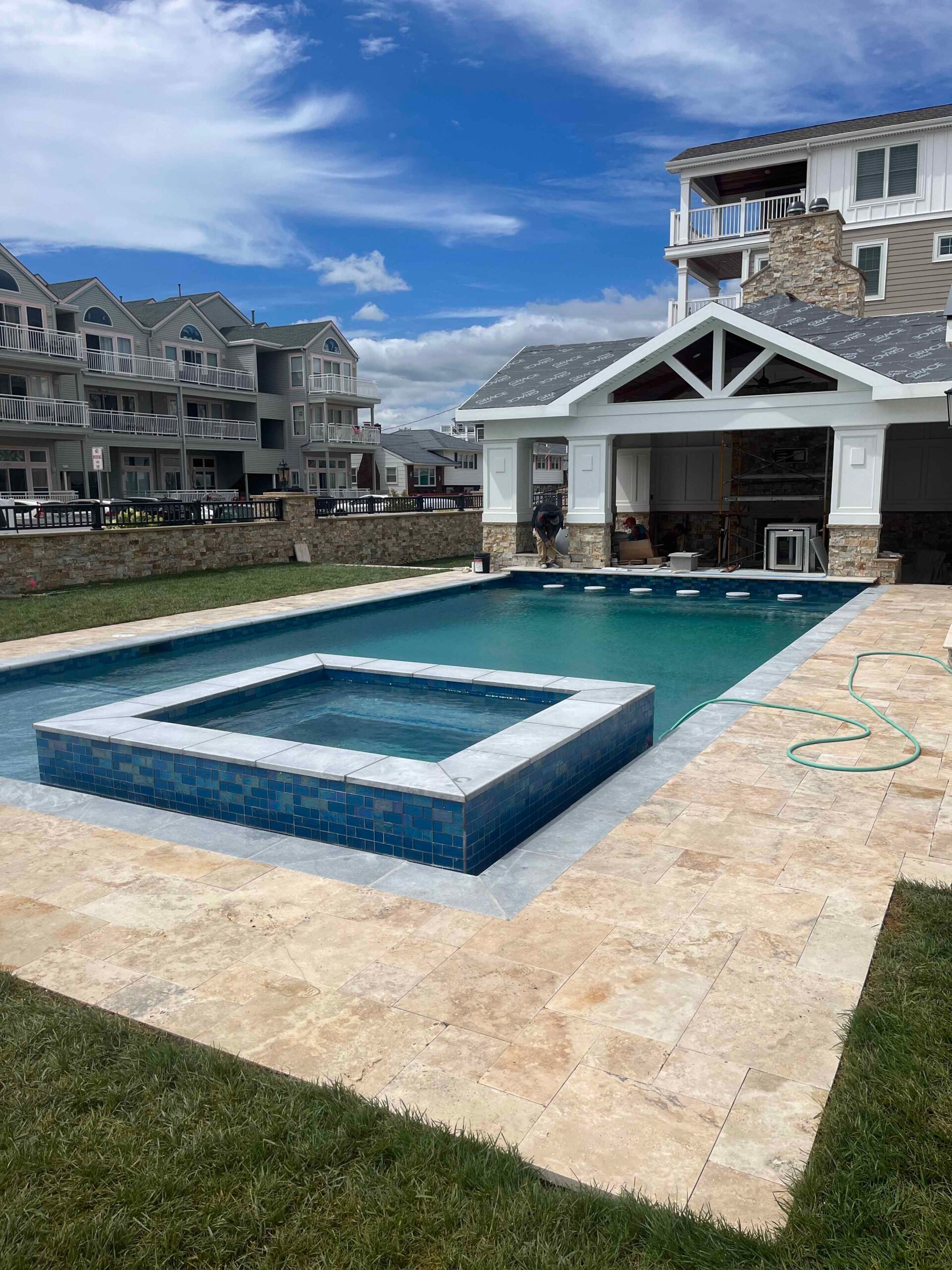 A rectangular in-ground pool with an attached square spa sits on a travertine paver surround. The spa is tiled in blue glass mosaic, a pool house with stone veneer columns stands in the background, and a green lawn borders the left edge of the patio.