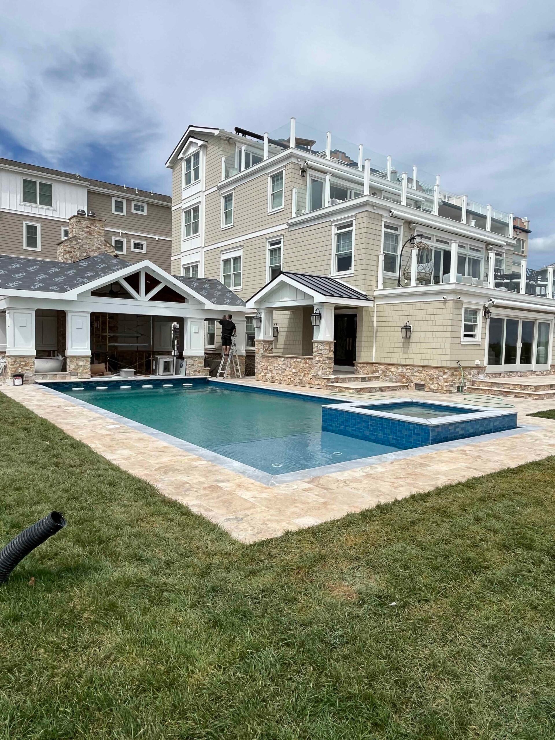 A rectangular in-ground pool with an attached spa sits on a travertine paver pool deck surrounding all sides. The lawn area adjacent to the patio shows recently laid sod. A pergola-covered outdoor living structure and a multi-story coastal home with stone 