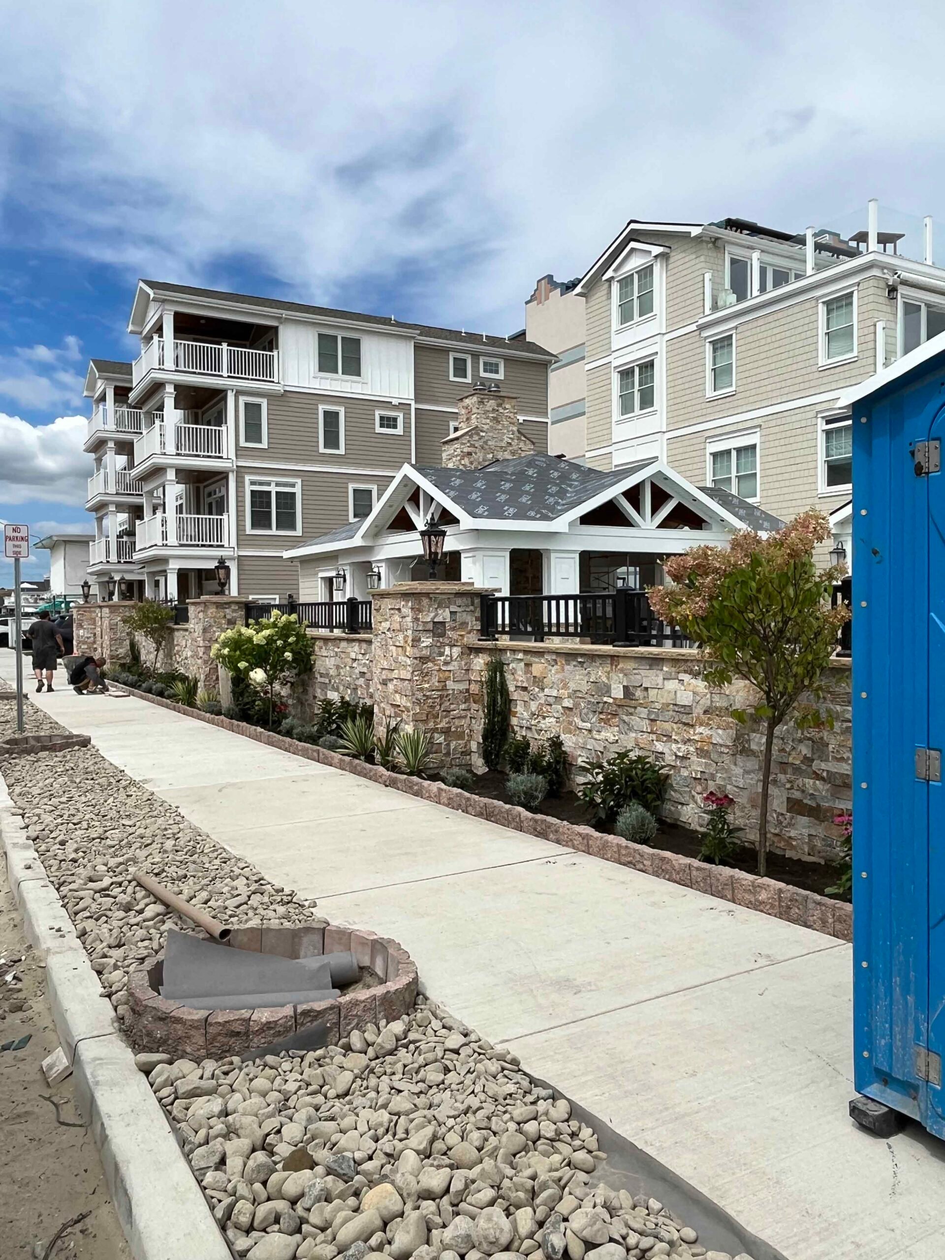 A completed natural stone retaining wall with stacked stone pillars borders a concrete sidewalk along a coastal commercial property. Planting beds behind the wall contain ornamental grasses, white flowering shrubs, small deciduous trees, and low succulents