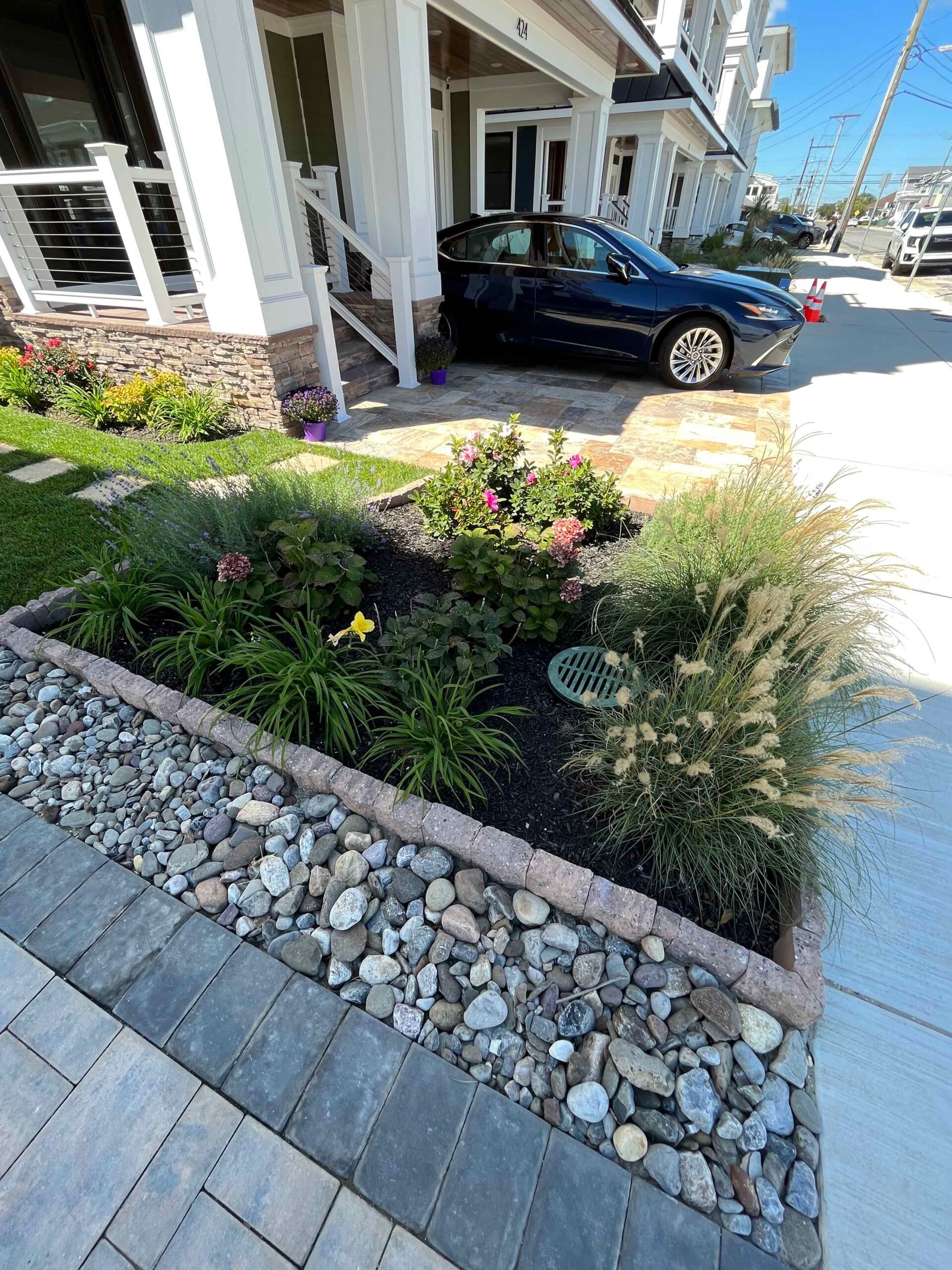 A front entry area features a concrete paver driveway with a raised planting bed bordered by tumbled concrete edging stones and a river rock dry swale. The bed contains ornamental grasses, flowering rose shrubs, and perennials mulched with dark black mulch