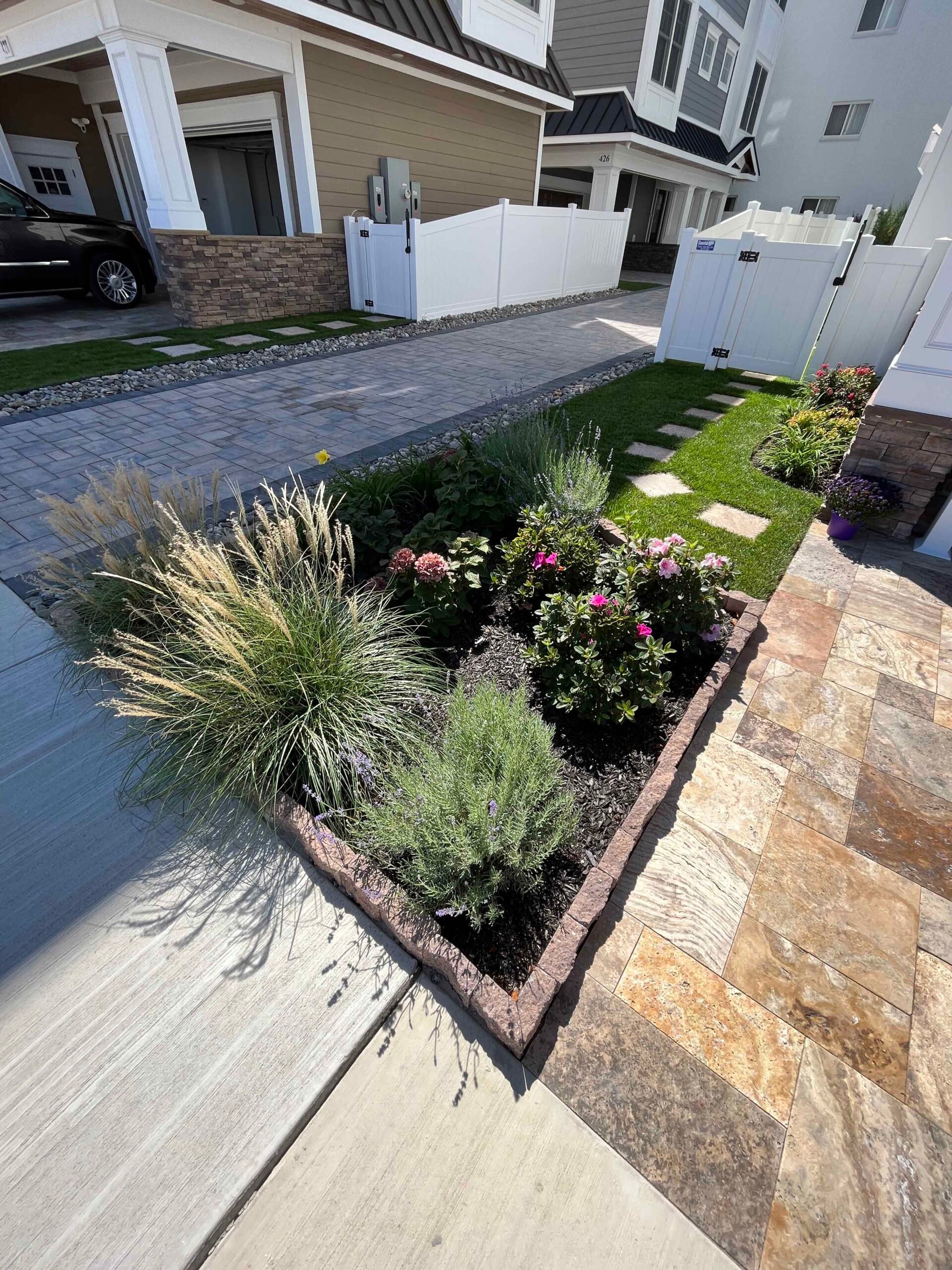 A gray concrete block paver driveway runs alongside a raised planting bed edged with brick border containing ornamental grasses, lavender or artemisia, and pink-flowering roses or knockout roses set in dark mulch. A natural stone tile walkway with a traver