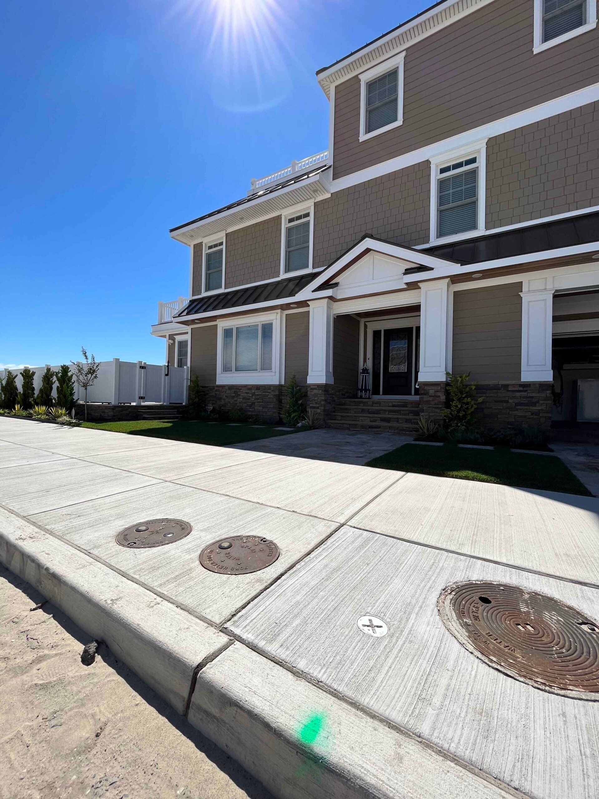 A freshly poured large concrete driveway leads to a two-story home with stone veneer foundation accents. Narrow planting strips along the front of the house contain young arborvitae, ornamental grasses, and low shrubs in freshly prepared beds. The lawn are