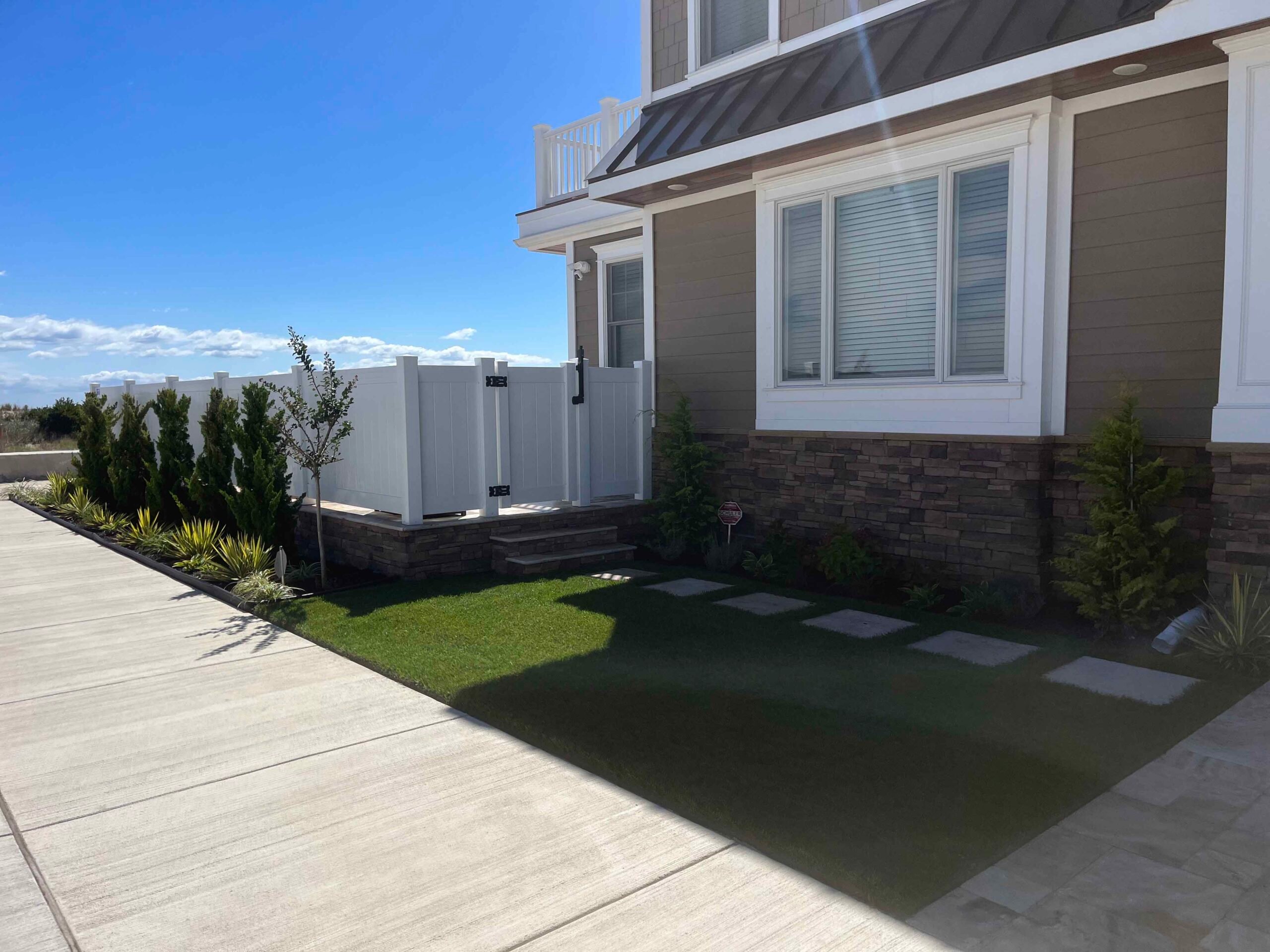A front yard features a small artificial turf lawn bordered by a concrete driveway and a planting bed with young arborvitae, ornamental grasses, and a small deciduous tree. Rectangular concrete stepping stones lead from the driveway to a white vinyl gate.
