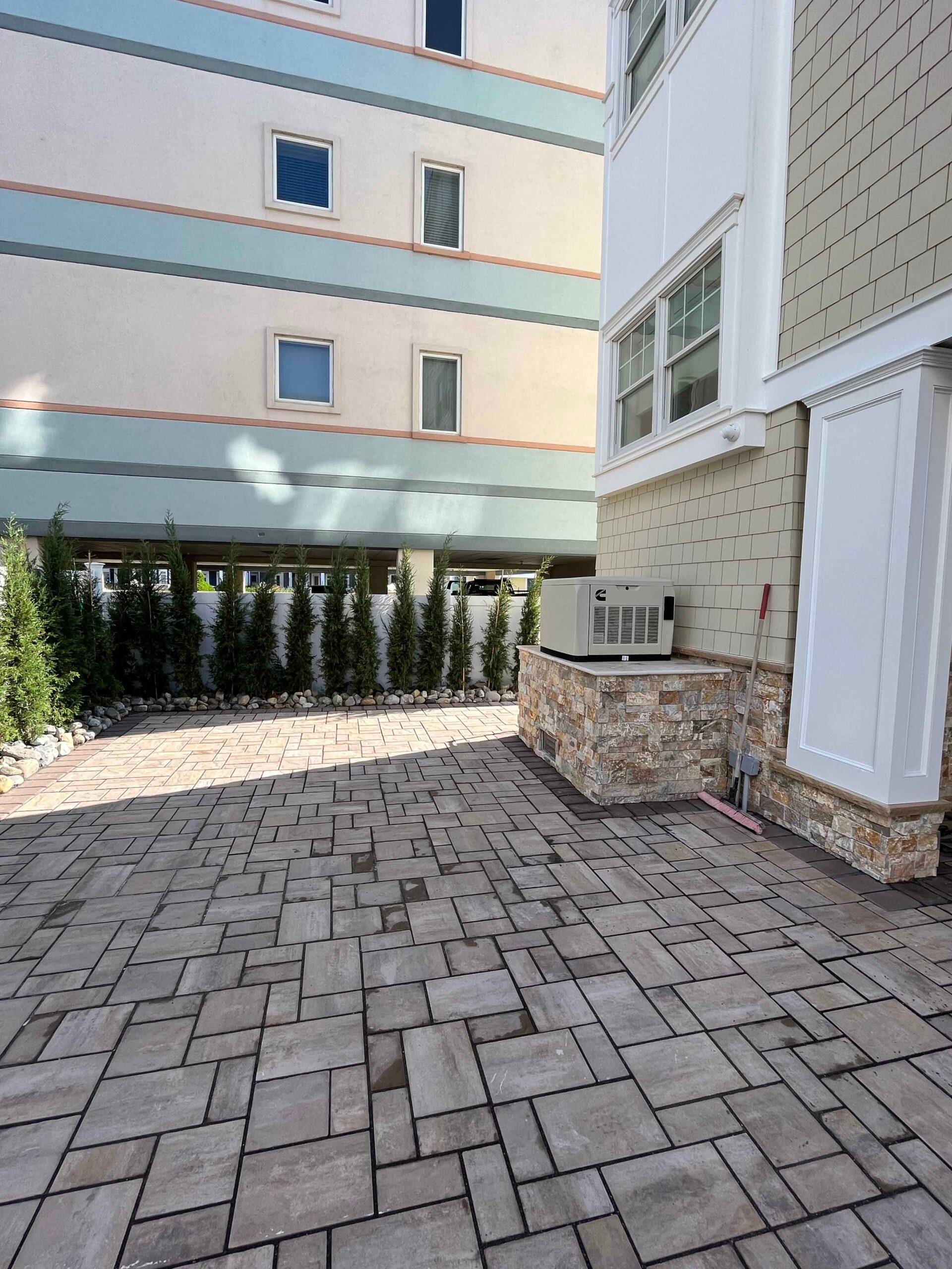 A large commercial courtyard paved with rectangular concrete pavers in a varied gray tone sits between two multi-story buildings. A row of upright arborvitae is planted along the far edge in a bed bordered by white river rock, providing a privacy screen.
