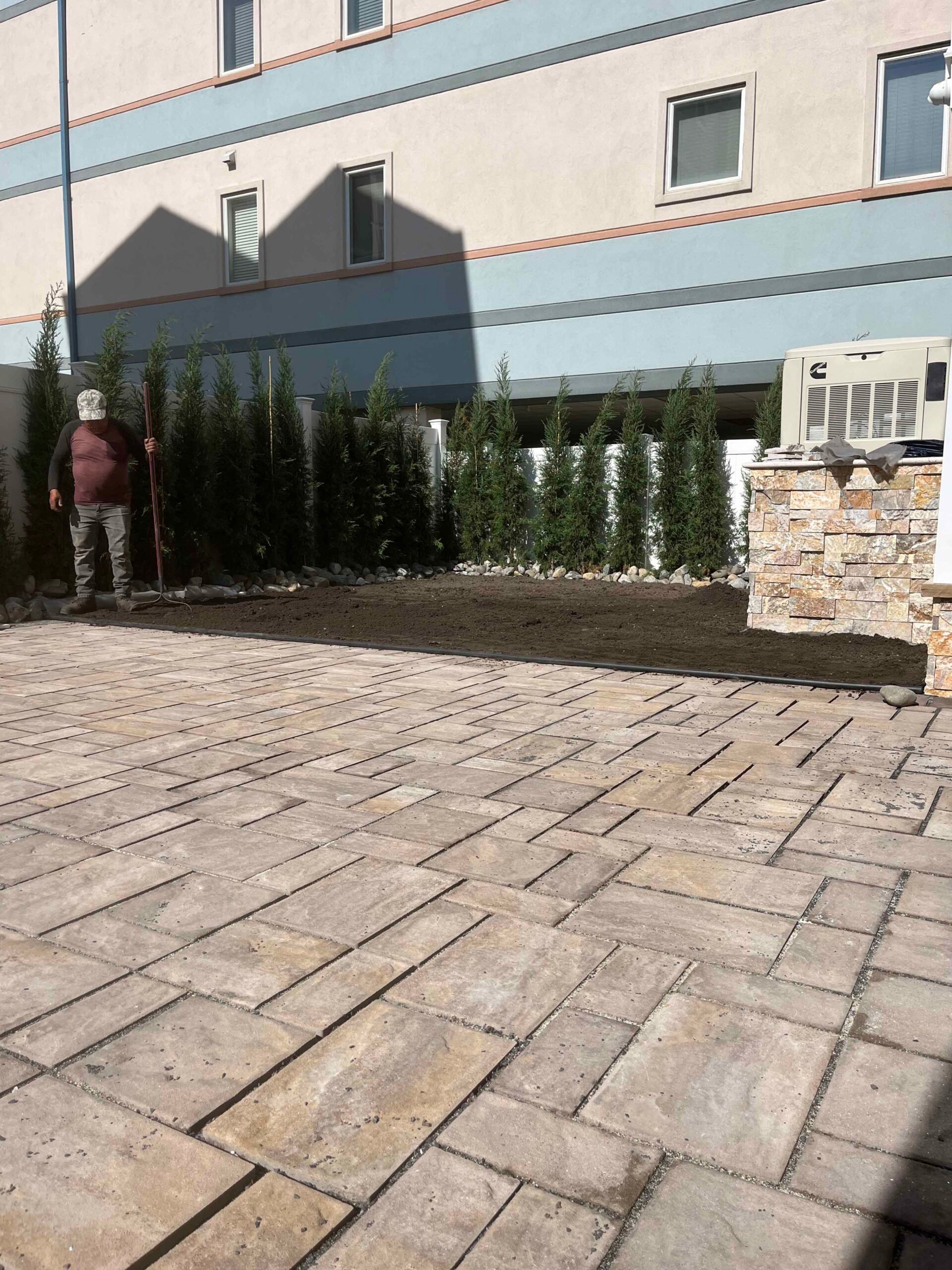 A wide commercial or residential patio surface laid with large-format tan travertine or concrete pavers occupies the foreground. Along the building foundation, a freshly graded planting bed contains a row of columnar evergreen trees, likely Italian cypress