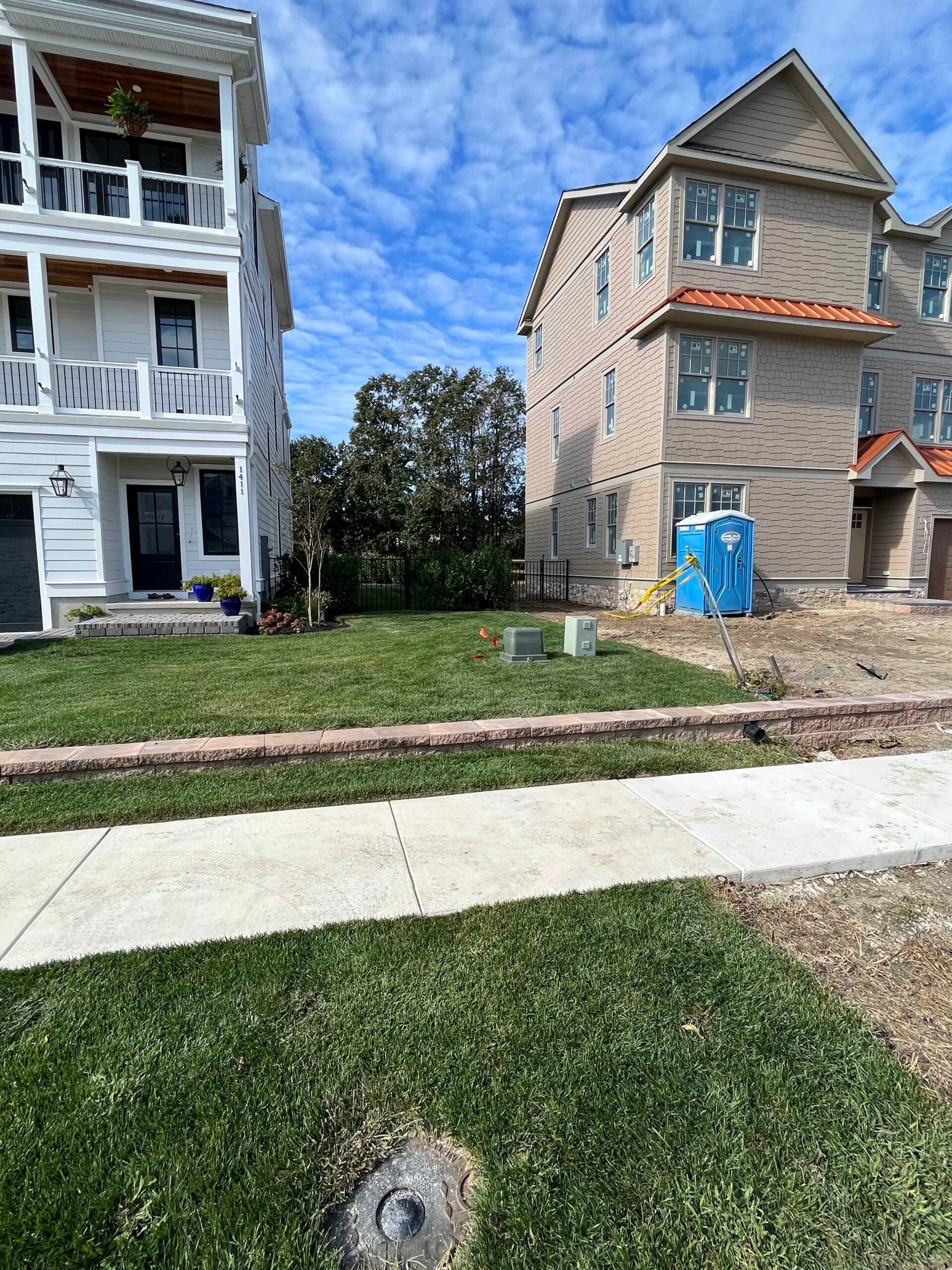 Newly installed sod covers the front yard between two multi-story homes, with a concrete sidewalk and paver curb border defining the lawn edge. The site shows active construction with a portable restroom and utility boxes visible, indicating a new landscap