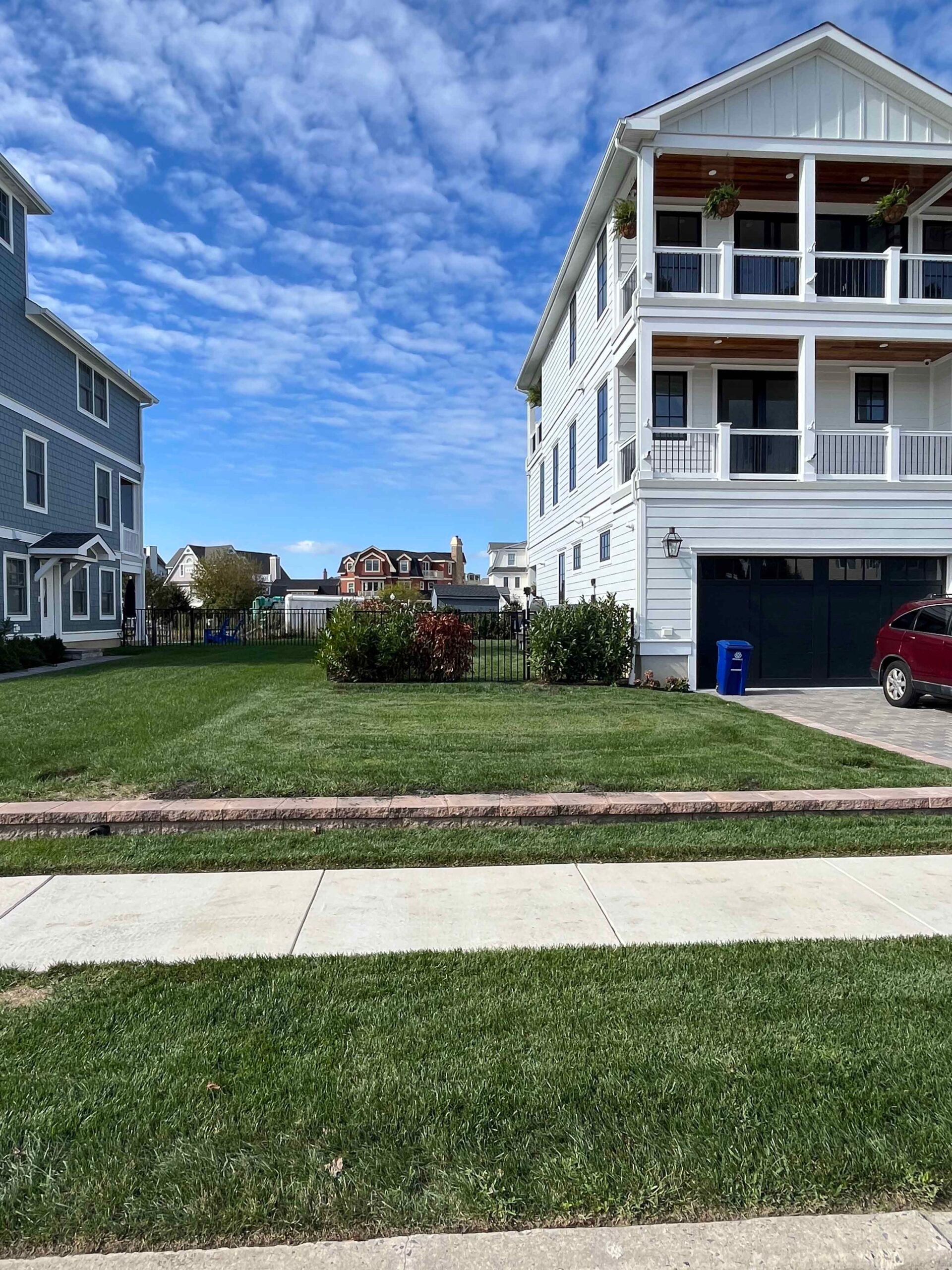 A freshly mowed residential front lawn with dense green turf stretches to the sidewalk in front of a multi-story white coastal home. Low shrubs including red-leafed and green varieties line the fence near the garage, and a paver or brick curb edge separate