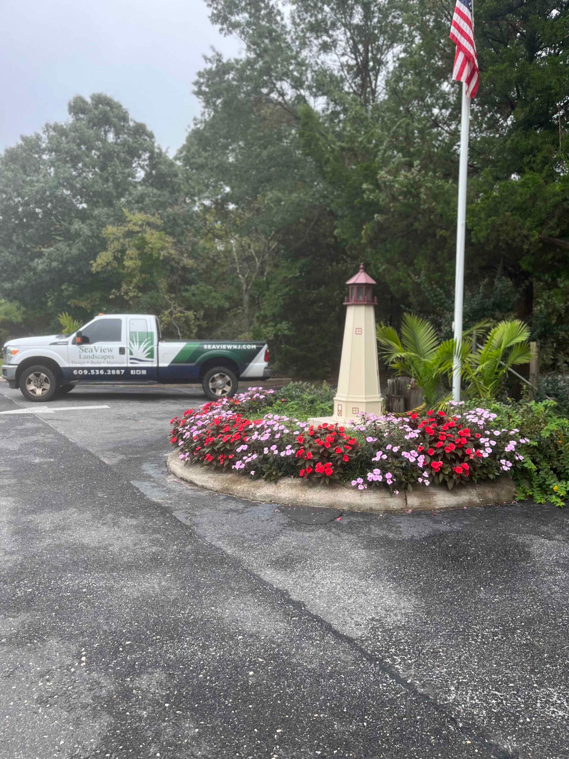 A rounded concrete-edged island planting bed contains dense red and pink impatiens surrounding a decorative wooden lighthouse ornament and flagpole. A SeaView Landscapes branded truck is visible in the background beside mature deciduous trees.