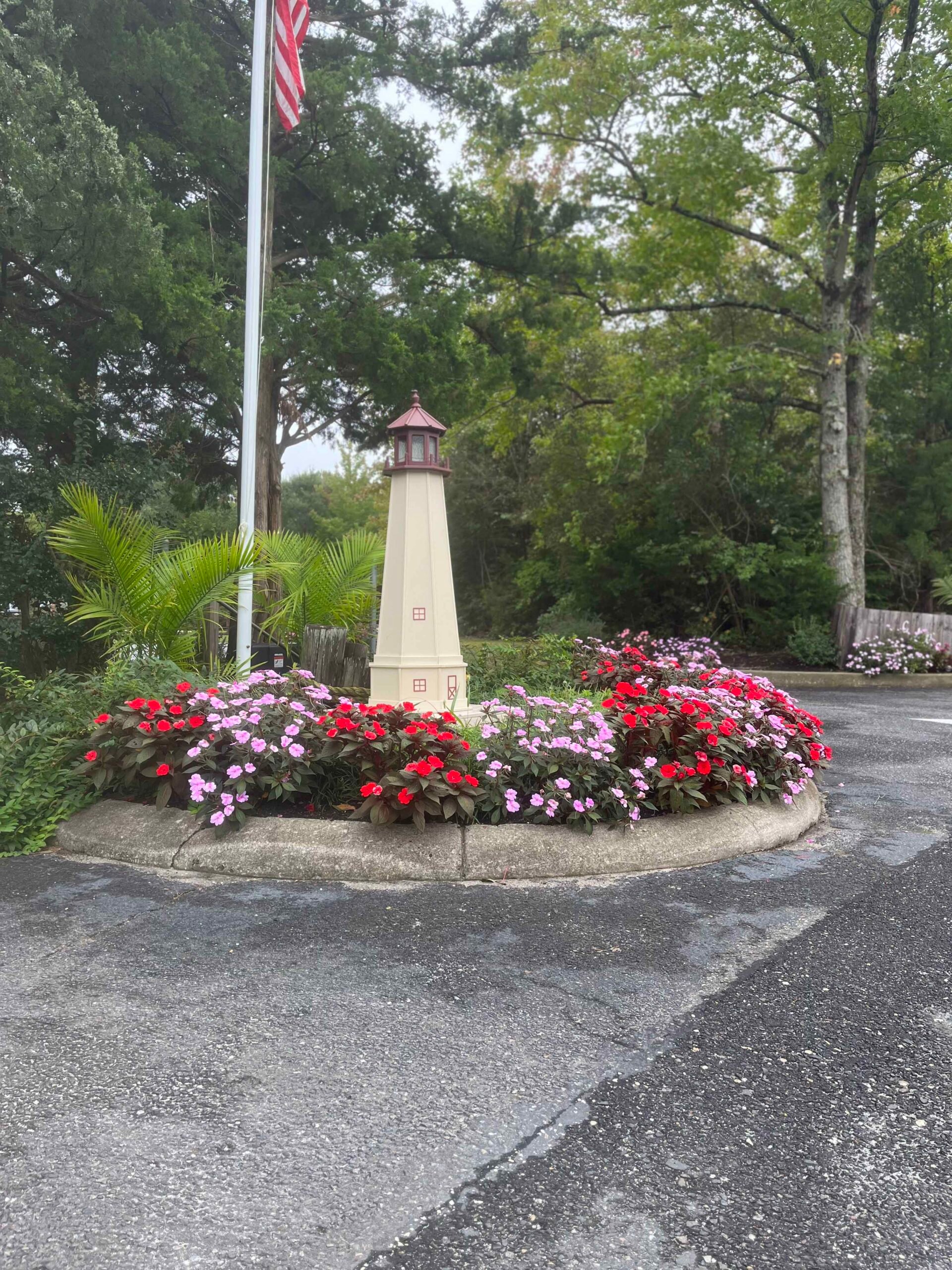 A close-up view of a rounded concrete-edged island bed planted densely with red and pink impatiens in full bloom surrounding a decorative painted wooden lighthouse. A flagpole and mature deciduous and palm-like trees fill the background.
