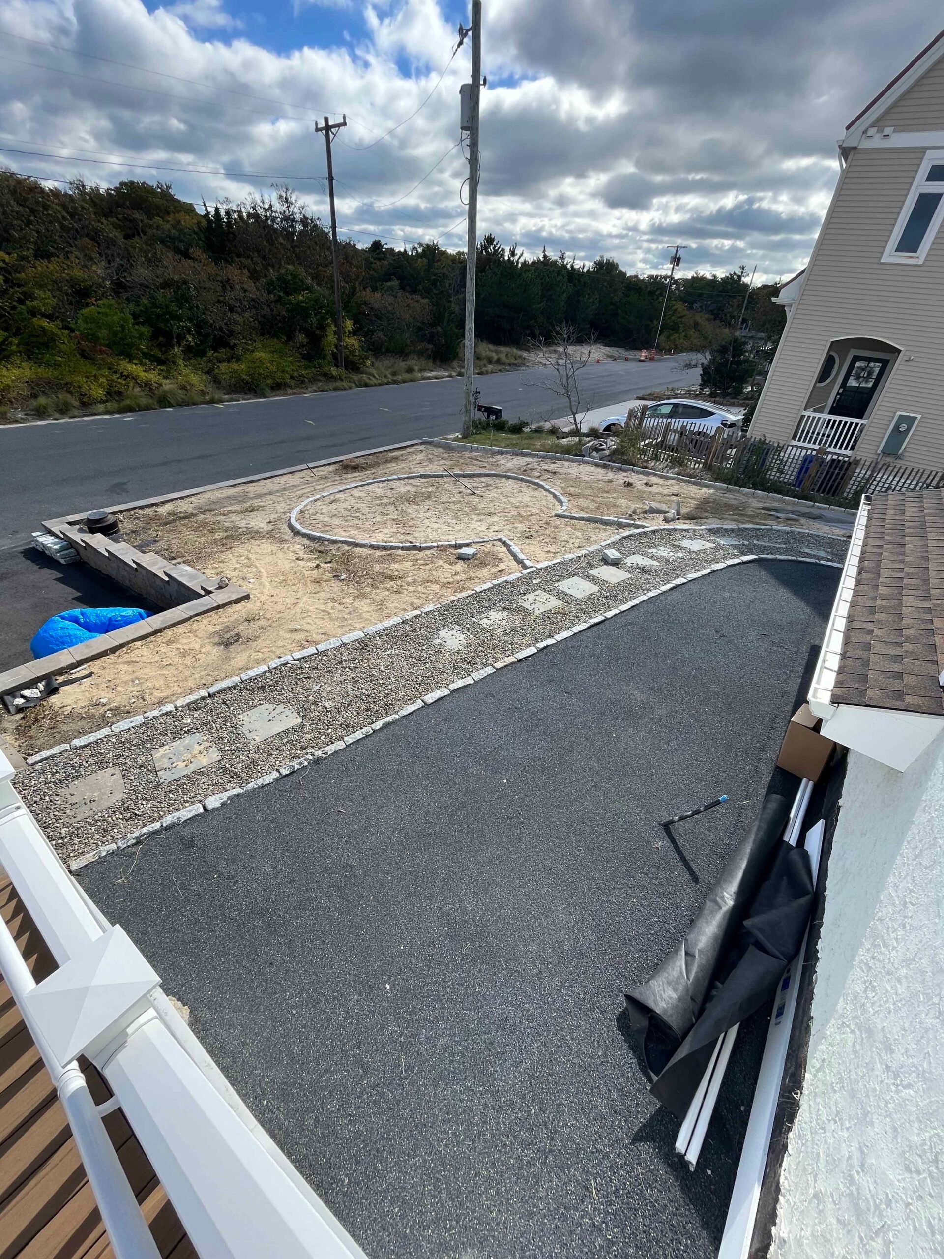 An aerial view of a residential property showing a new asphalt driveway alongside a partially constructed planting bed layout marked out in gravel and pavers on bare soil. The surrounding area has natural scrub vegetation and the project is in an active co