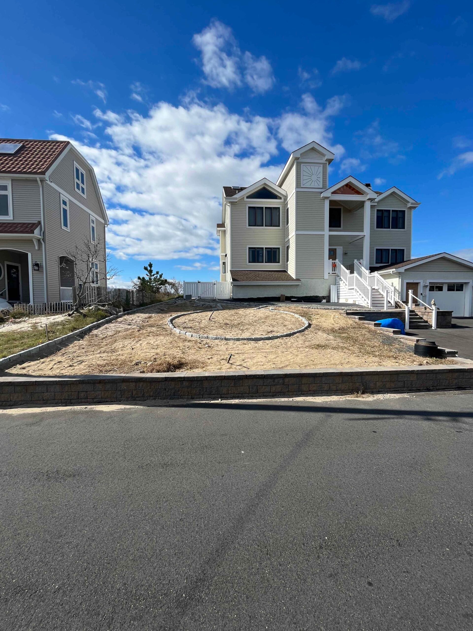 A front yard shows a freshly graded and compacted sandy soil area with a circular paver pattern laid out in the center, likely marking a future planting bed or focal point feature. A segmental block retaining wall runs along the street edge. The yard is in