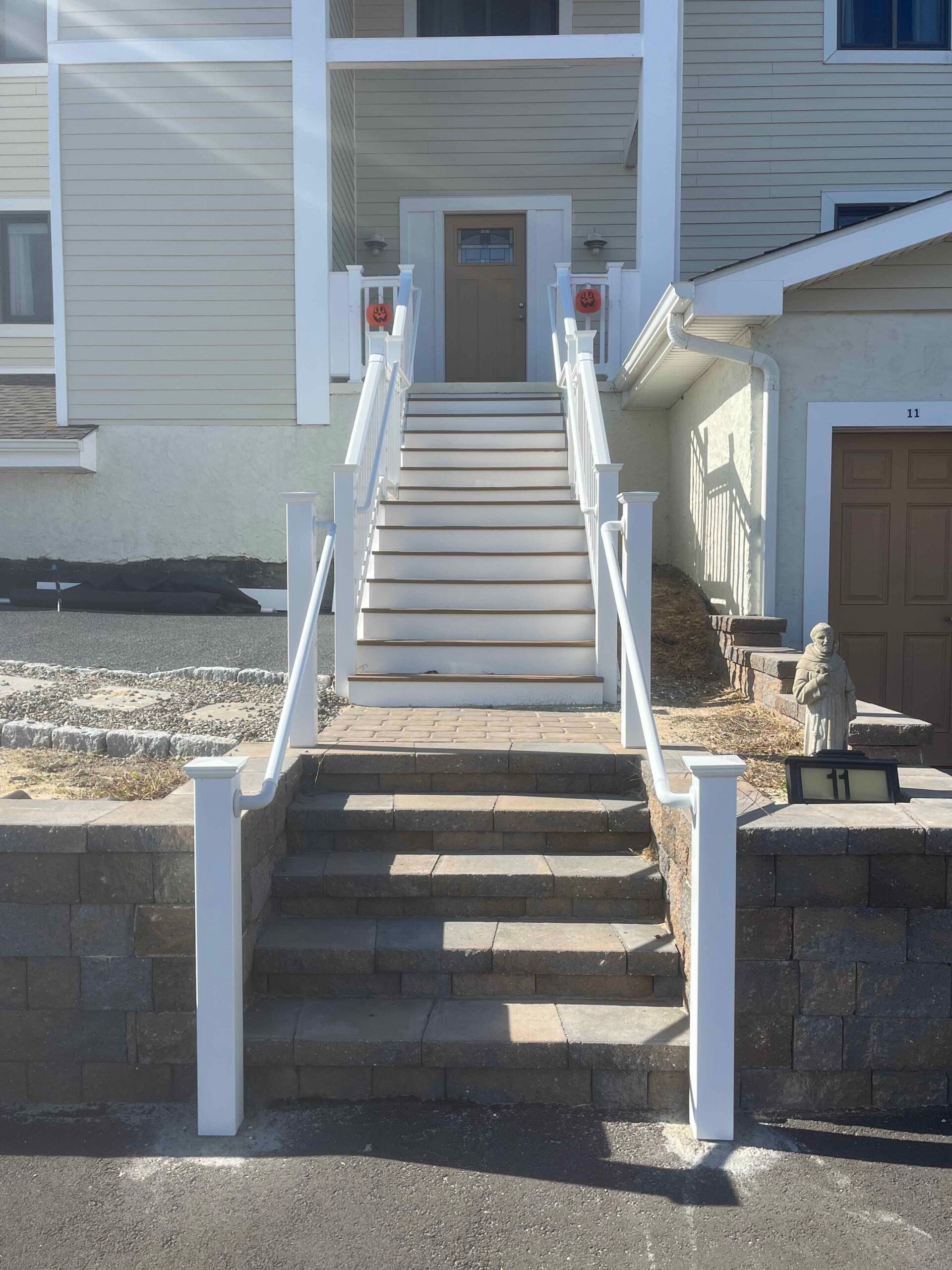 A multi-level concrete paver block retaining wall flanks a front entry staircase with concrete treads and white vinyl railings; the surrounding ground is bare soil with scattered stone, indicating a landscape construction or seasonal cleanup phase.