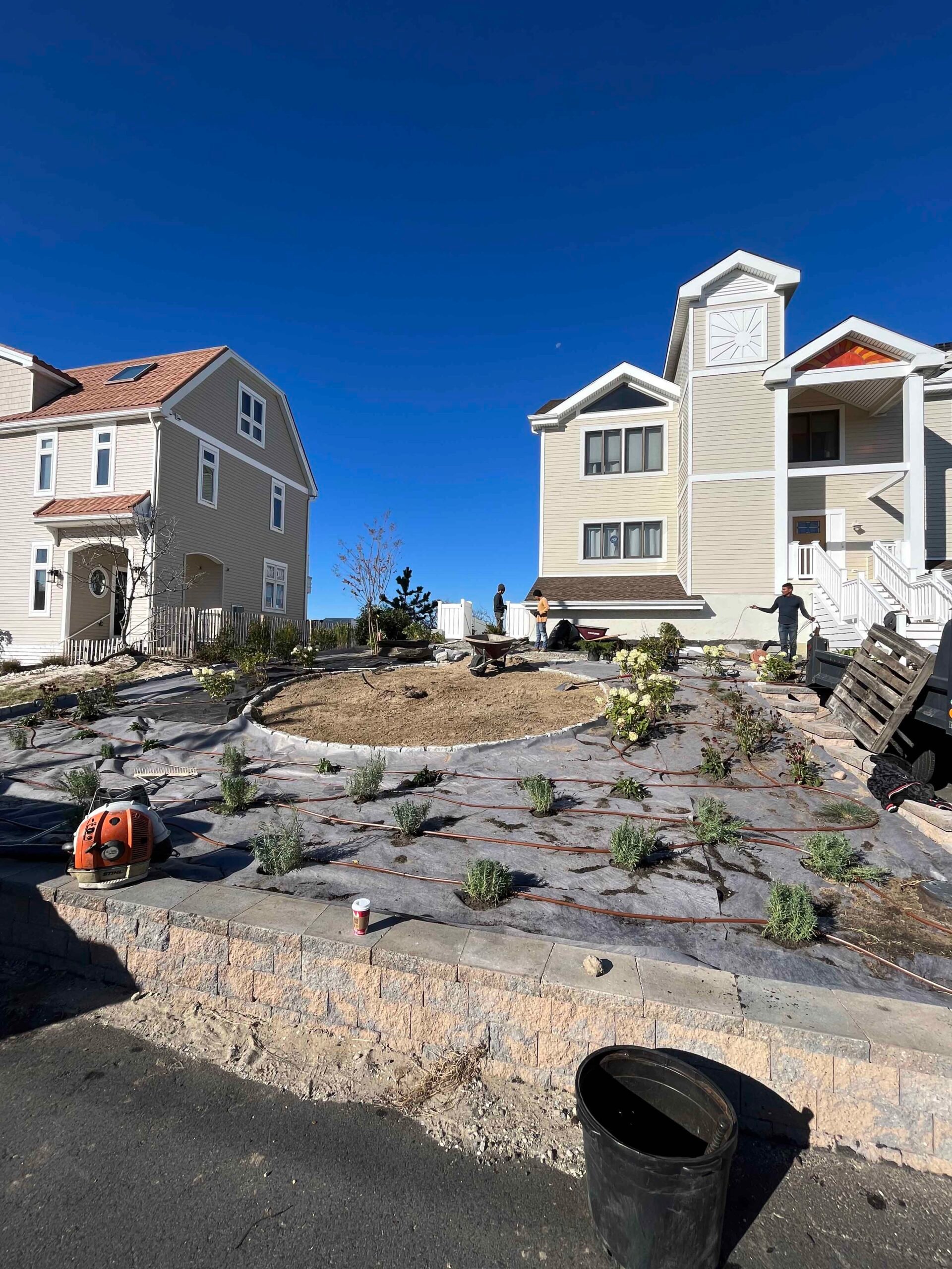Crew members install young shrubs and perennials across large prepared planting areas covered in landscape fabric, with drip irrigation lines visible. A circular island bed is taking shape in the center, bordered by a segmental concrete block retaining wal