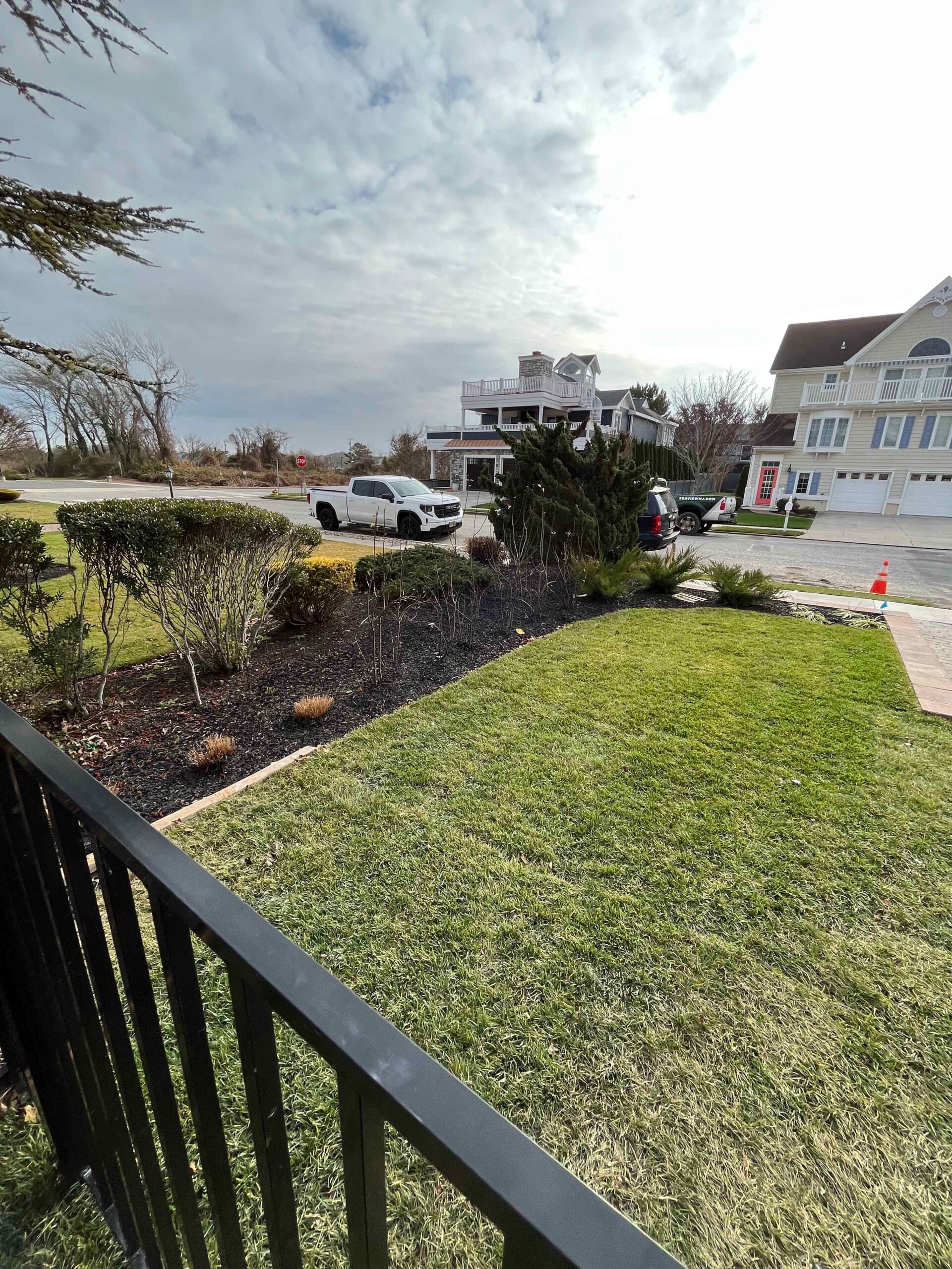 A front yard planting bed filled with dark mulch contains rounded evergreen shrubs, ornamental grasses, and low ground cover plantings along a manicured green lawn. The bed runs along a concrete paver border with a metal railing visible in the foreground, 