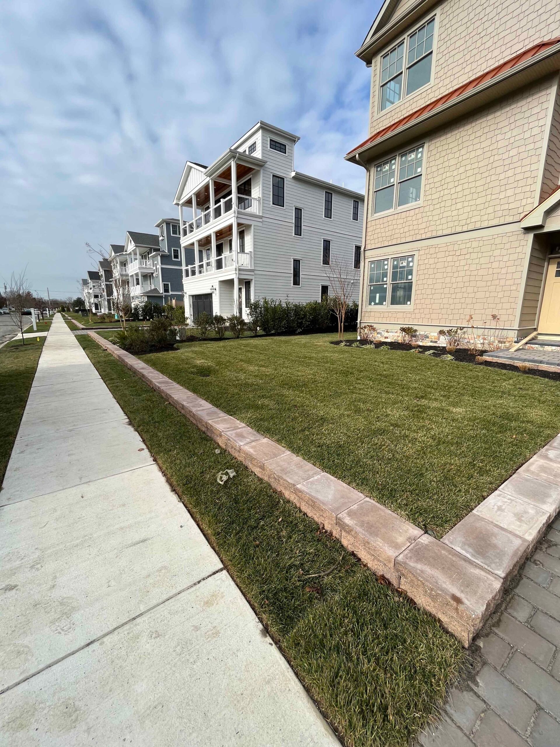 A freshly sodded front lawn runs along a concrete sidewalk, bordered by a low natural stone paver retaining edge. Newly planted mixed shrubs line the foundation of a coastal-style multi-story home in the background.