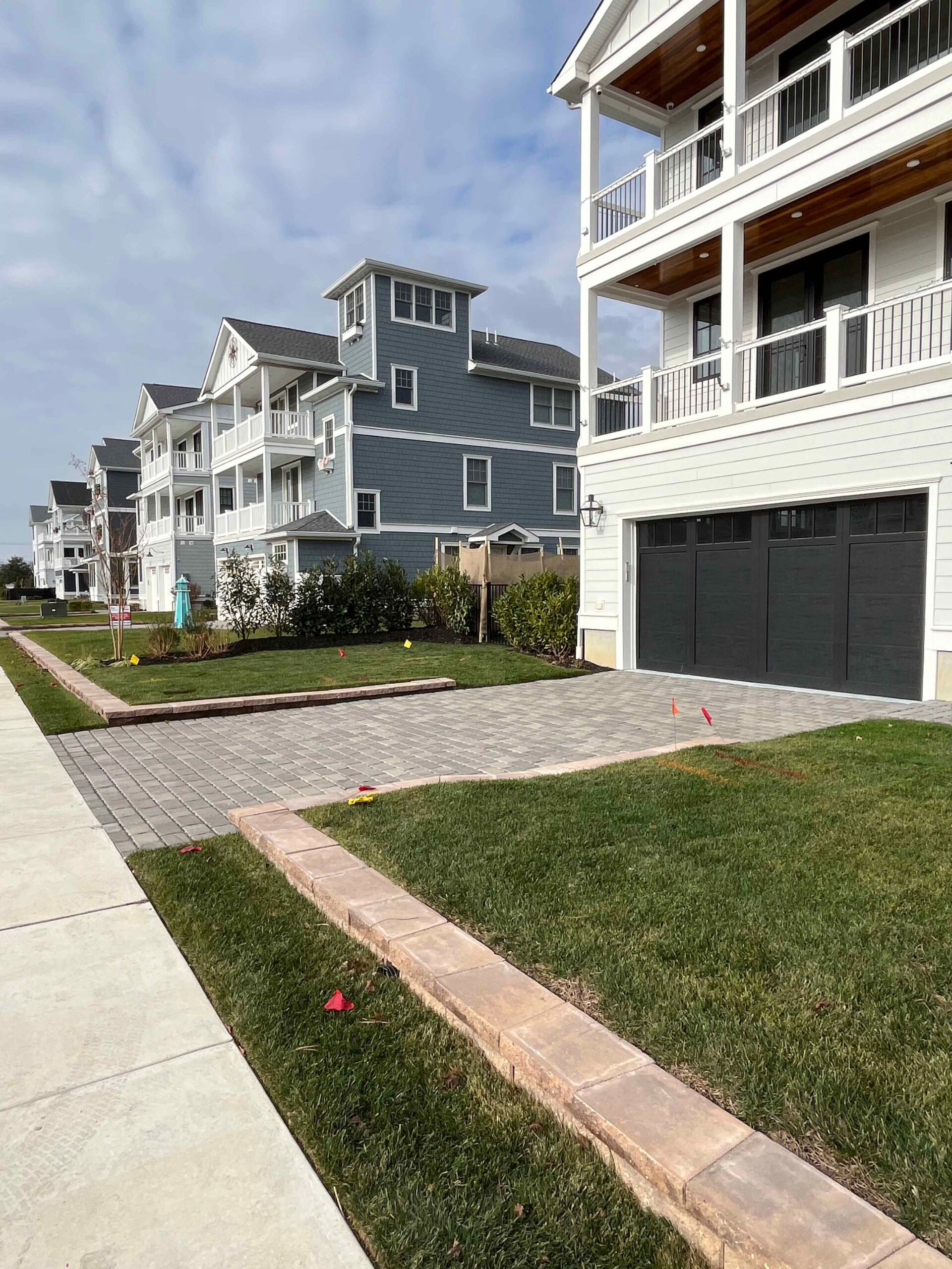 A newly installed concrete paver driveway apron in a running bond pattern leads to a two-car garage, flanked by freshly laid sod lawn strips bordered by a soldier-course brick curb. The setting is a coastal residential neighborhood with multi-story homes v