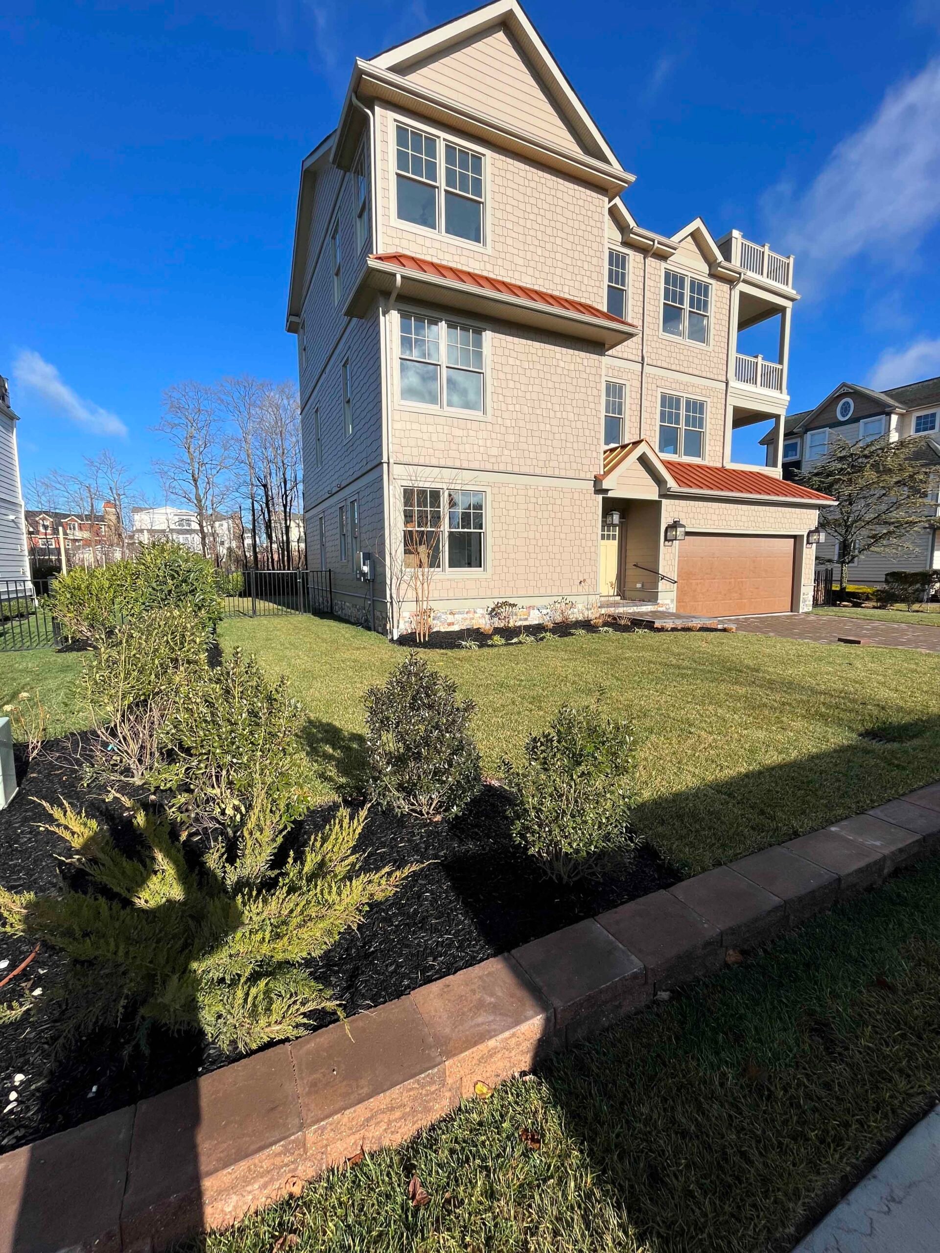 A newly installed front yard planting bed features low evergreen shrubs and ornamental grasses set in fresh black mulch, bordered by a brick paver retaining edge along a manicured lawn.