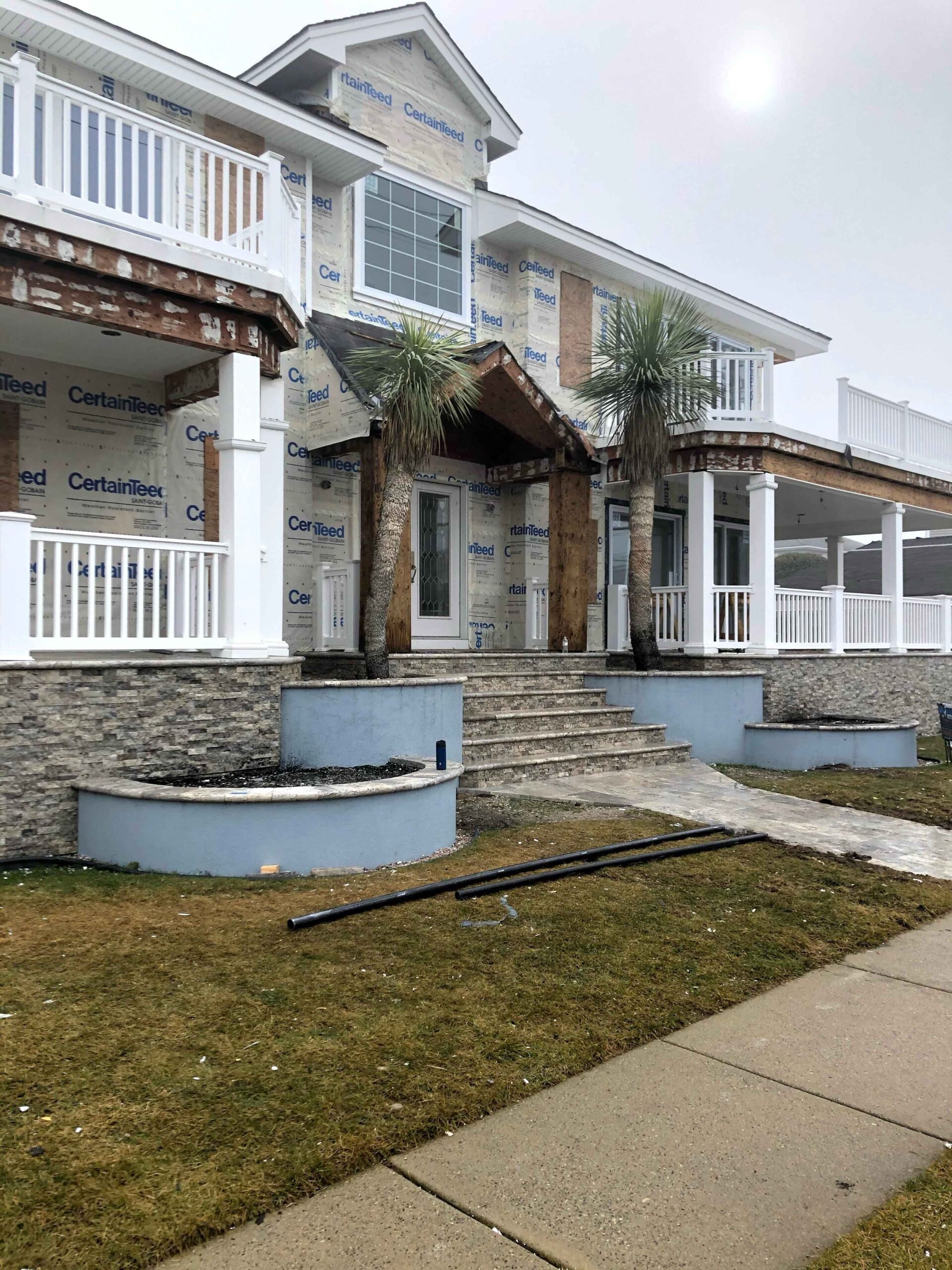 A residential property under construction shows two curved raised planting beds with stone veneer bases flanking a set of concrete entry steps. Two windmill palms are planted in the beds on bare soil with black irrigation pipe visible on the ground. The la