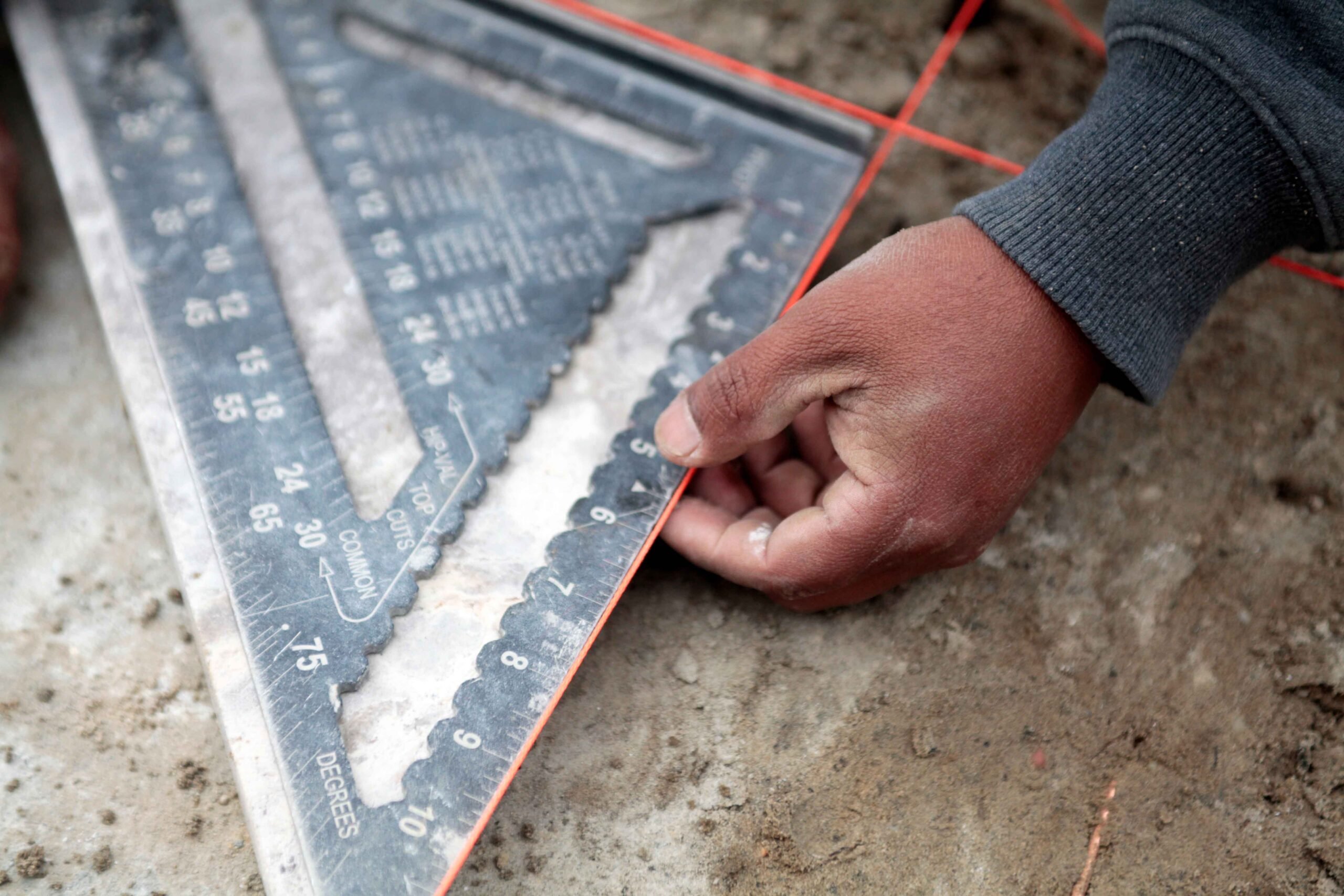 A worker uses a speed square and orange chalk line to measure and mark angles on a concrete or stone surface during hardscape installation. The surface appears to be bare ground or a slab in preparation for paving work.