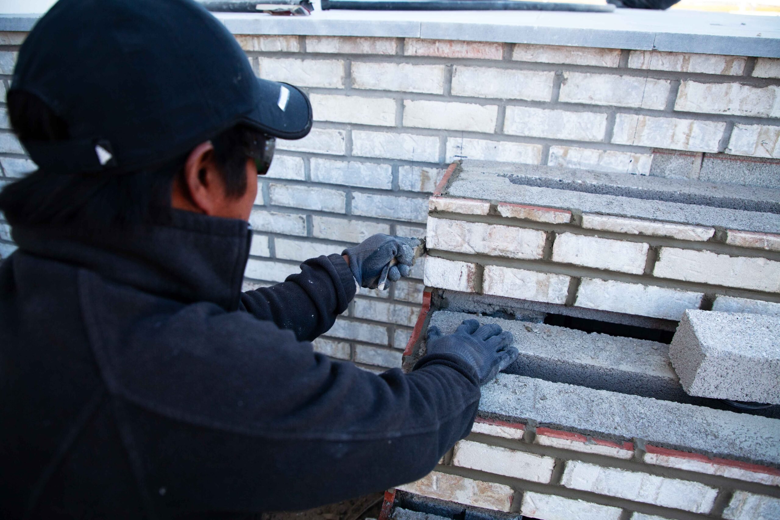 A worker applies mortar while laying white and gray brick courses on a retaining wall, with concrete block backing visible in the open cavity. The wall is mid-construction with a level line guide visible along the brick face.