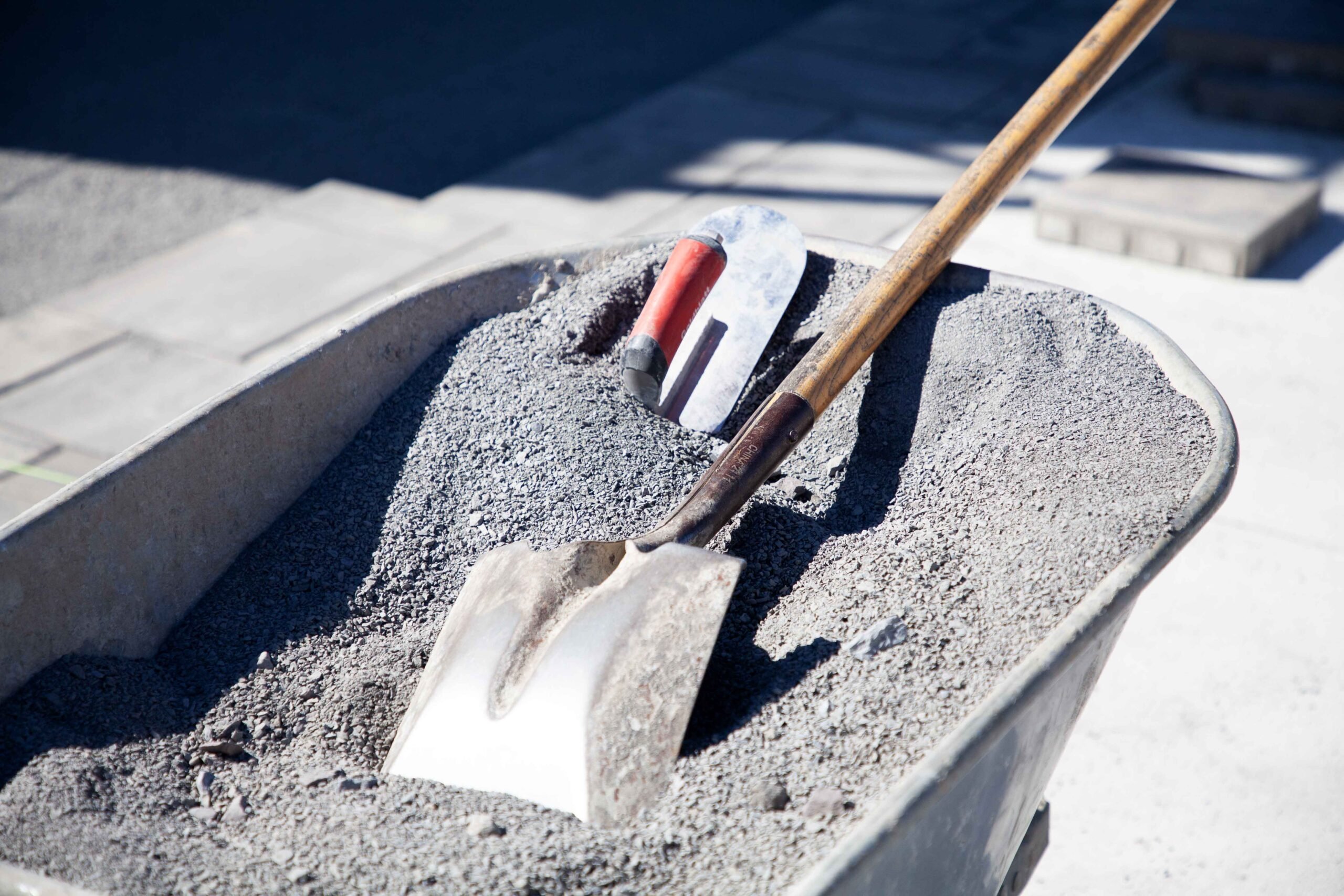 A metal wheelbarrow filled with crushed gravel aggregate sits on a concrete surface next to partially laid concrete pavers, with mason's tools including a flat shovel and pointing trowel visible in the aggregate.