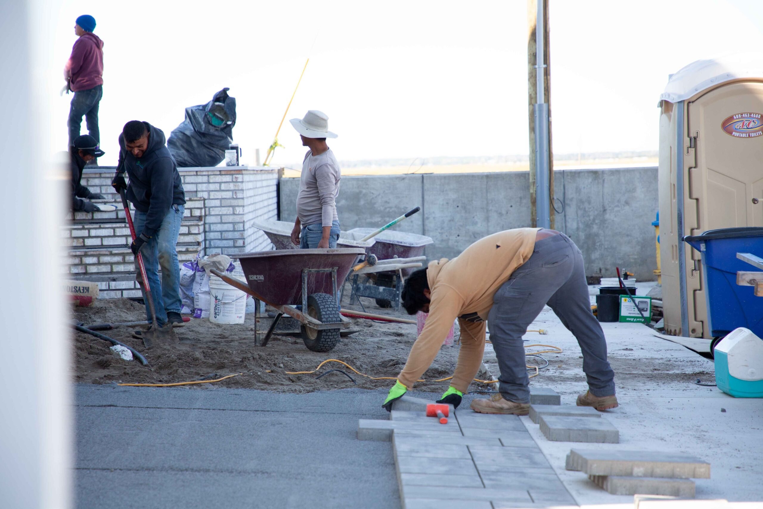 A crew of four workers installs concrete pavers at a construction site. Sand base is being prepared, a wheelbarrow holds material, and cut pavers are staged in the foreground next to a low white block retaining wall structure.