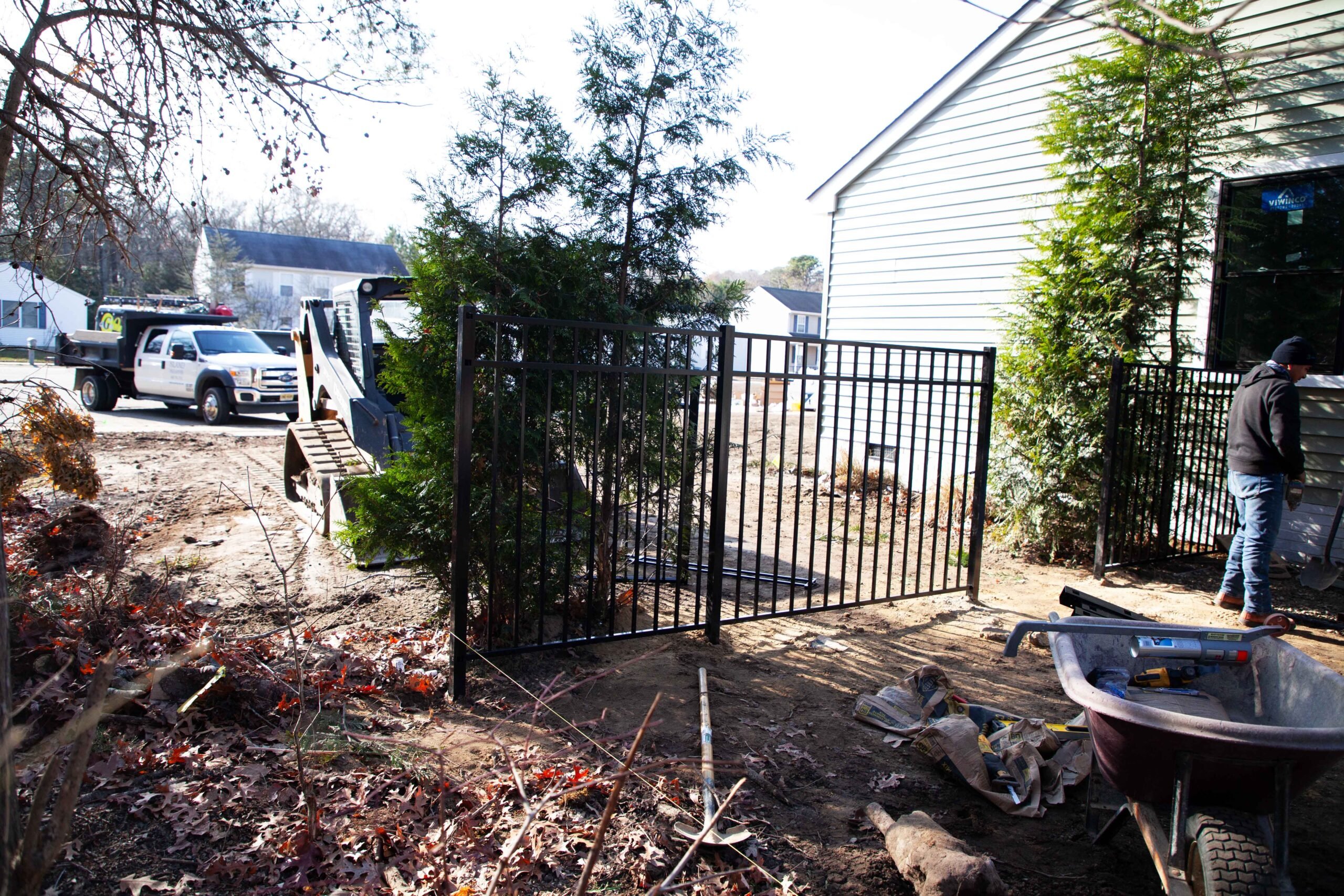 Two workers install a black steel spear-top fence gate at the side of a residential property. Arborvitae trees are planted along the fence line on bare, disturbed soil. A skid steer loader is visible in the background, indicating an active landscaping and 