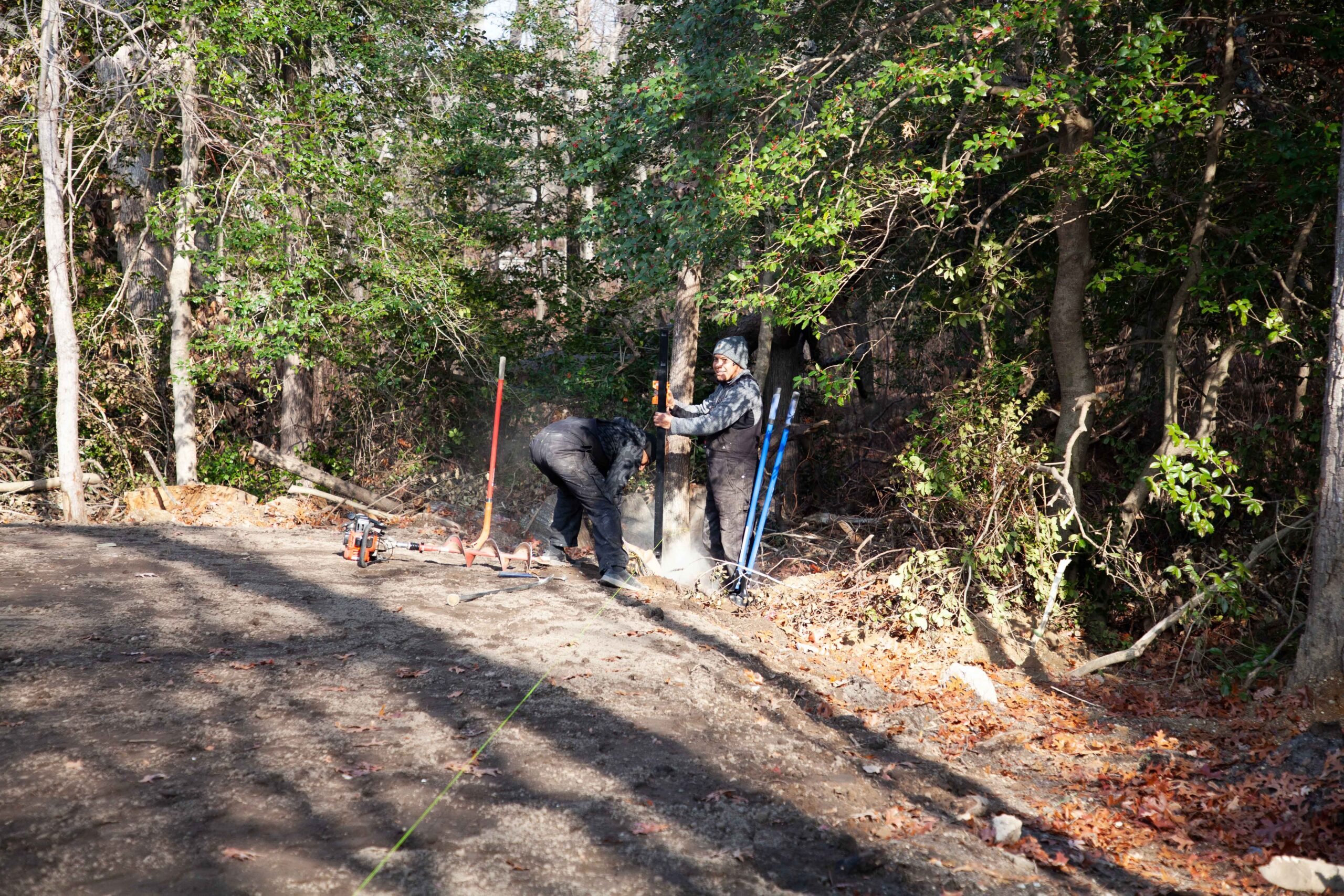 Two workers set a fence post along the edge of a wooded area with a mix of evergreen and deciduous trees. The ground is compacted gravel or packed earth, with leaf litter present at the tree line. The scene appears to be a late fall installation based on f