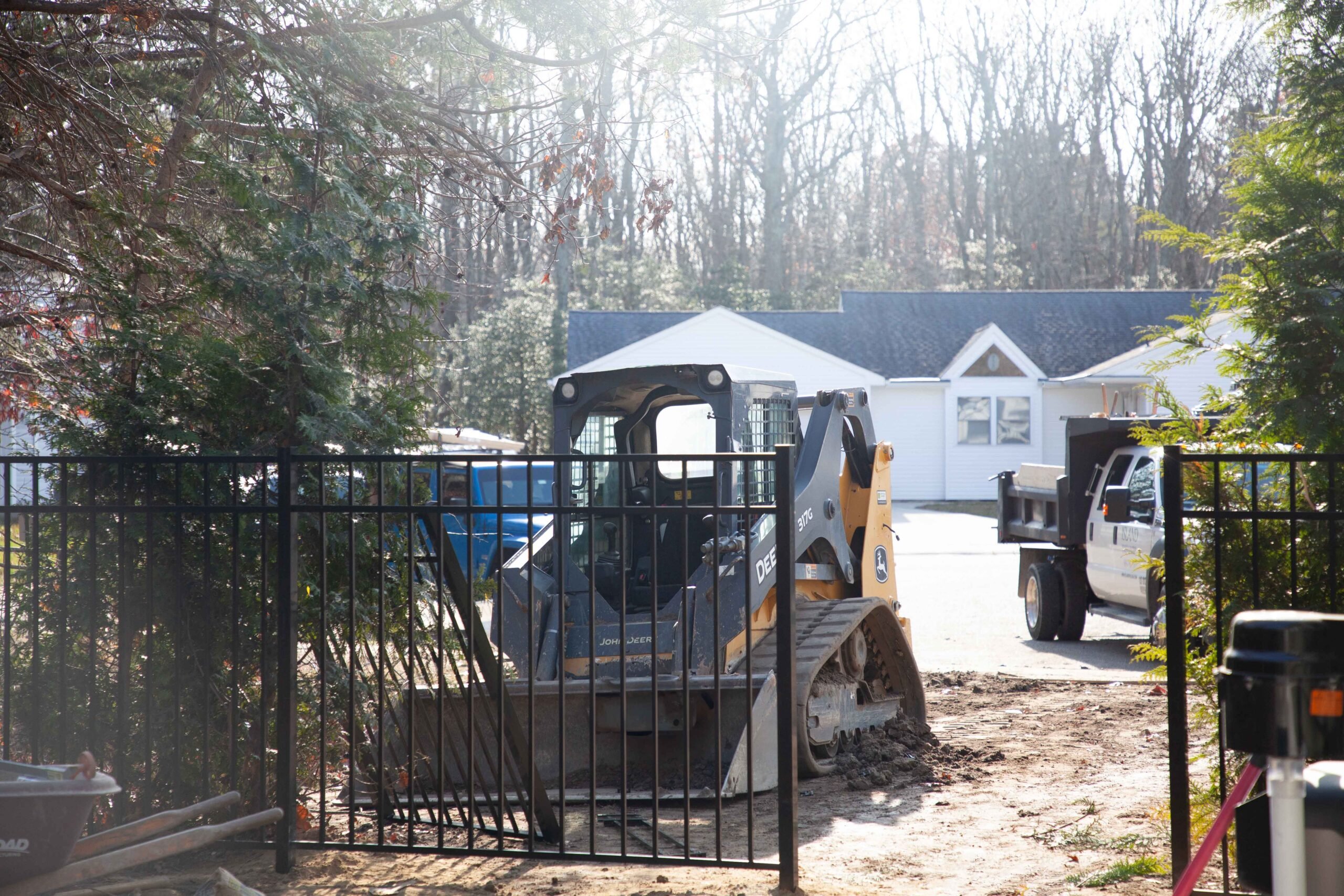 A John Deere skid steer loader operates inside a black aluminum fence line, moving soil during a site clearing or grading phase. Evergreen trees including what appears to be Eastern red cedar or arborvitae are visible to the left. A dump truck is parked in