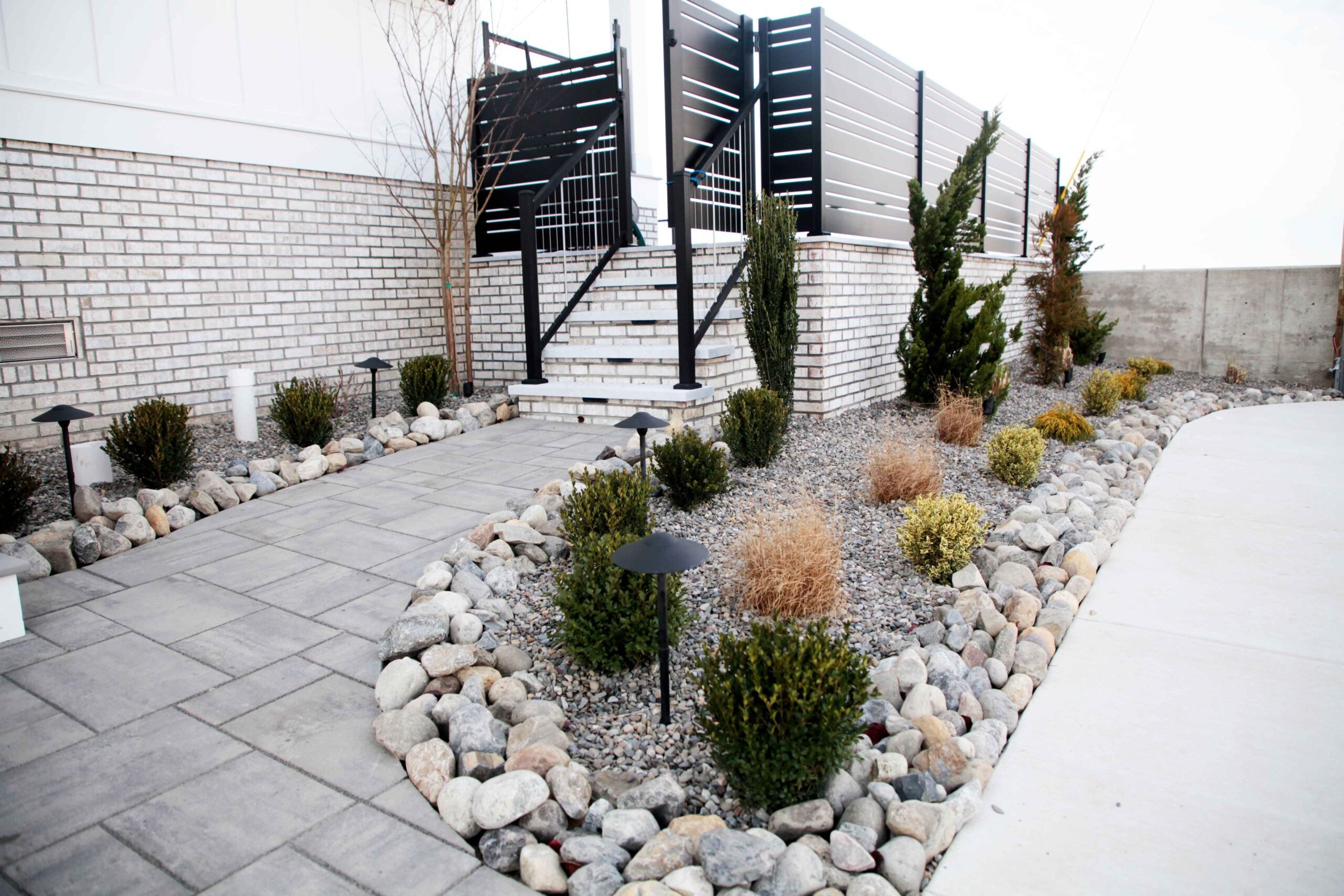 A concrete paver patio borders a river rock planting bed edged with rounded boulders, planted with columnar evergreens, dwarf rounded shrubs, and ornamental grasses; black low-voltage path lights are installed throughout the bed along a white brick wall wi