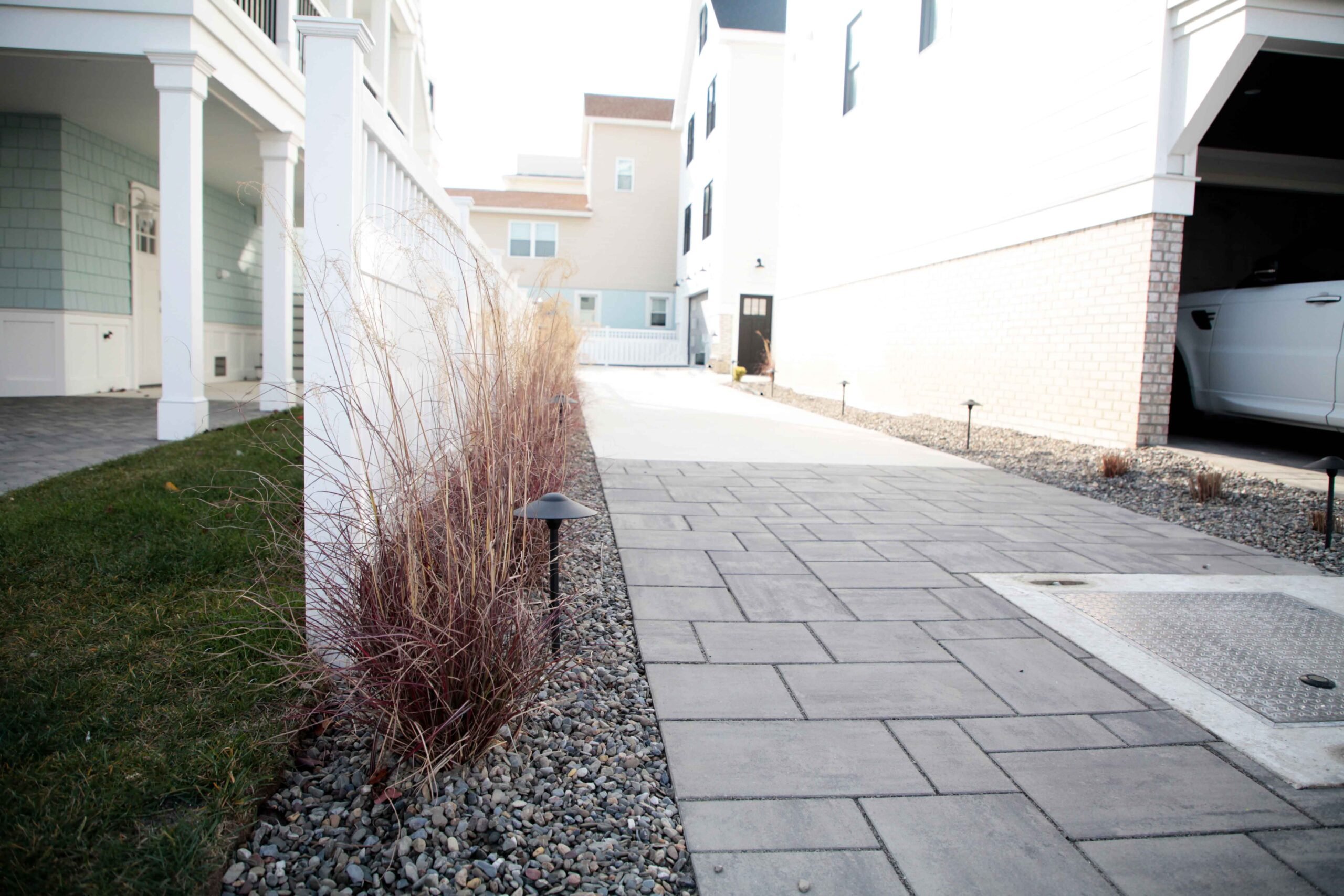 A concrete paver driveway runs between two buildings alongside a planting bed of reddish ornamental grasses growing in pea gravel, with low-voltage black path lighting fixtures installed at the bed edge.